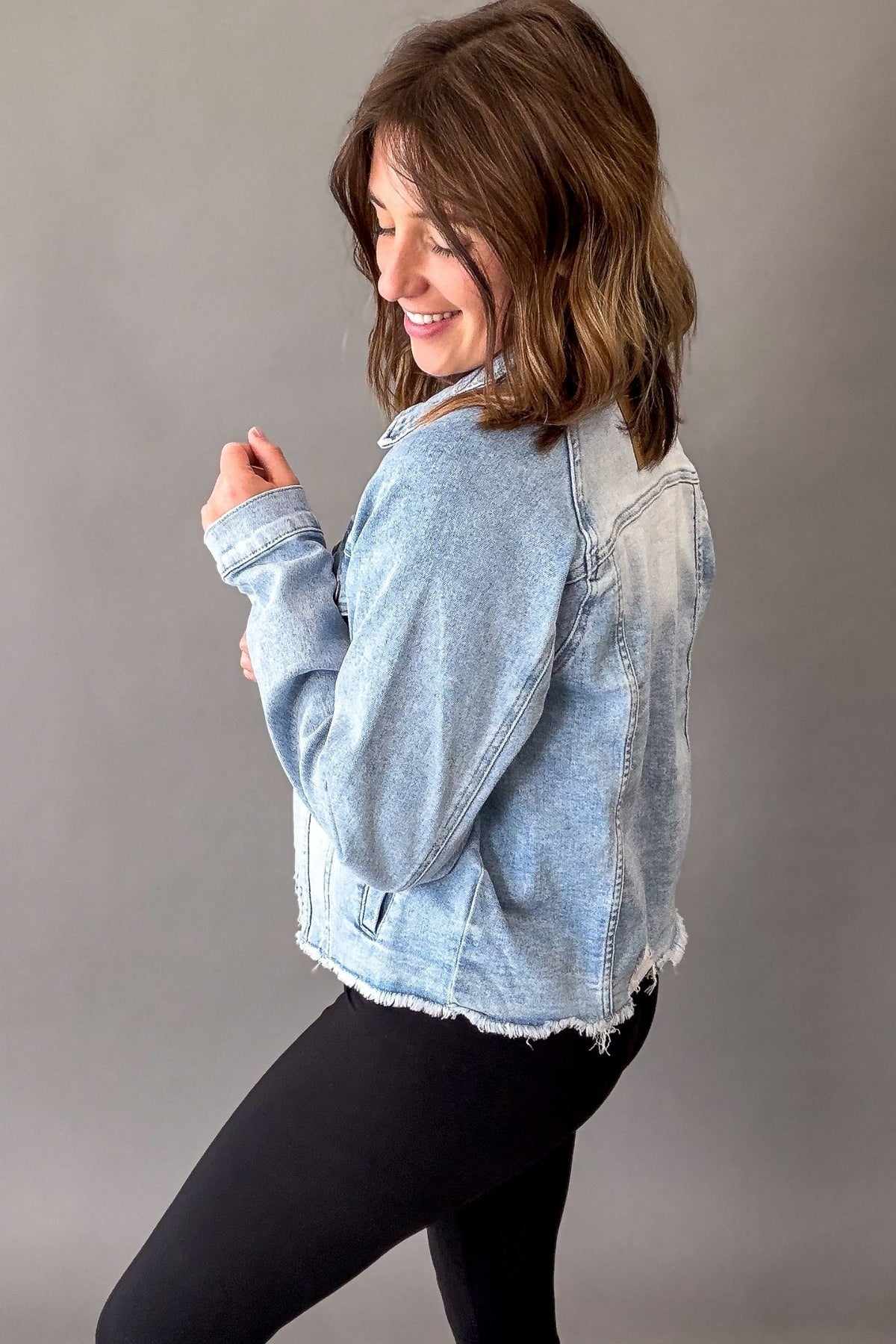 A woman wearing a light-wash blue denim jacket with raw, frayed hems and black leggings, posing against a solid gray studio background.