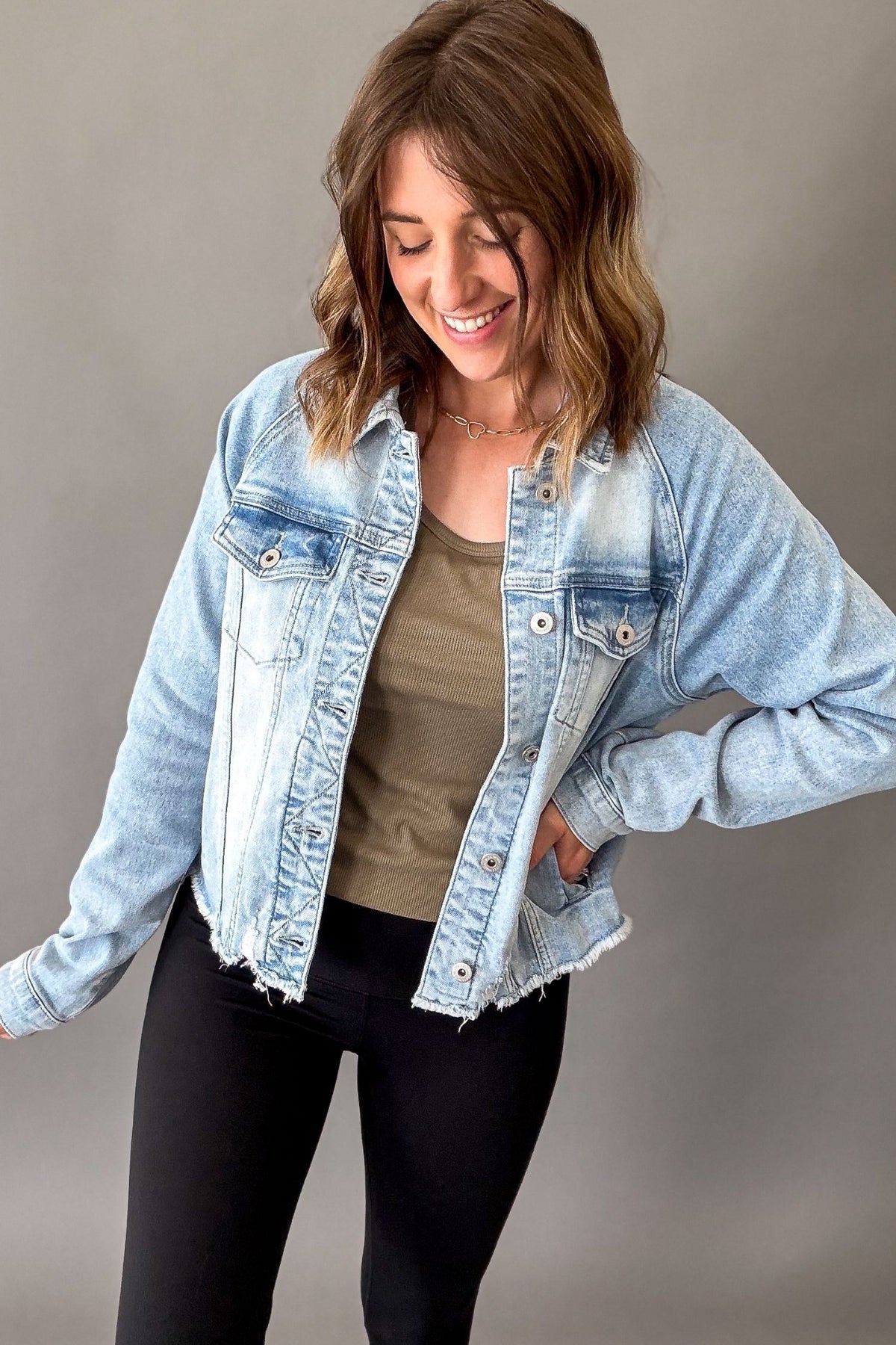 A woman wearing a light-wash blue denim jacket over a taupe tank top and black pants, posing against a neutral gray studio background.