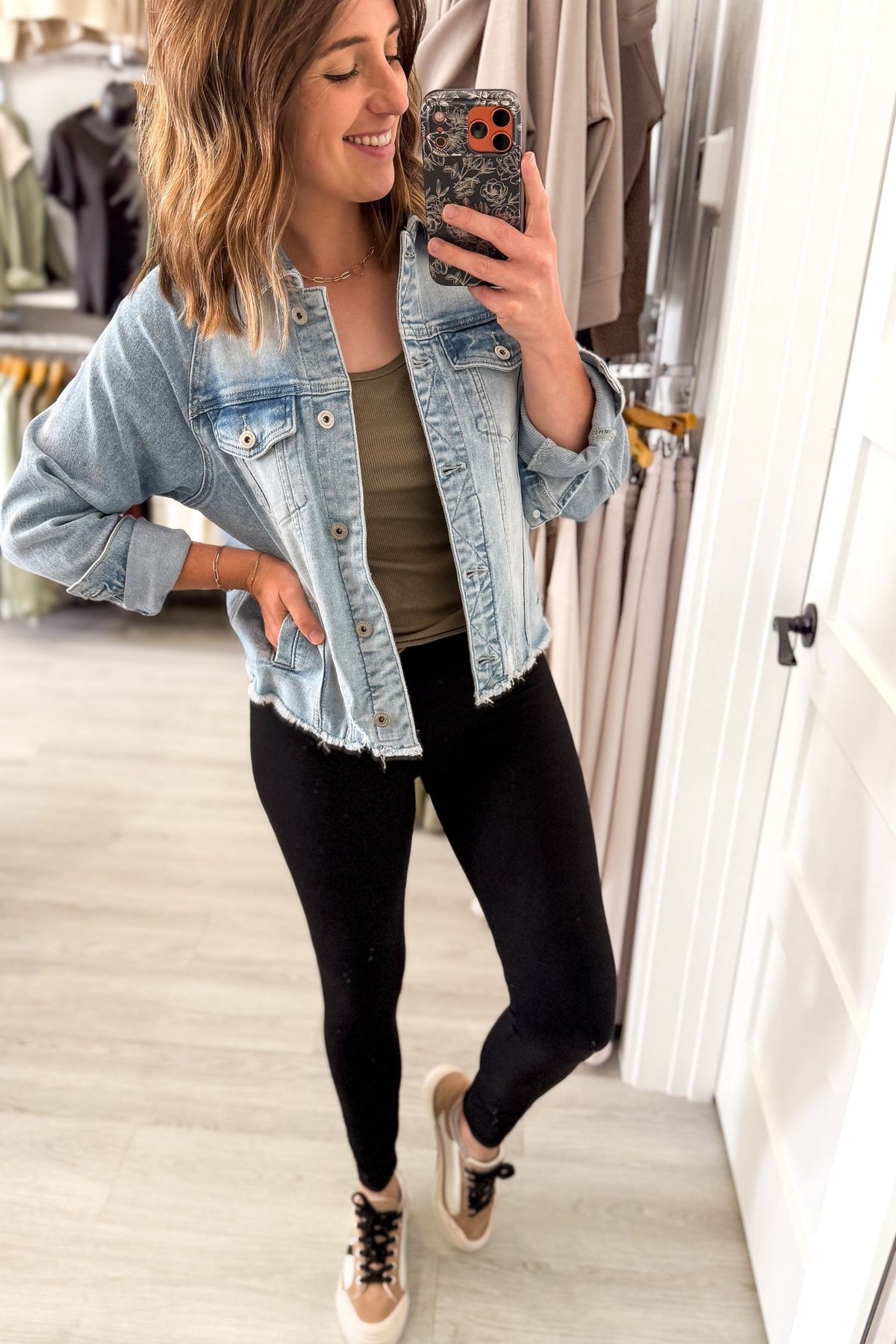 A woman takes a mirror selfie in a clothing store, wearing a light-wash blue denim jacket over an olive green tank top, black leggings, and beige sneakers.