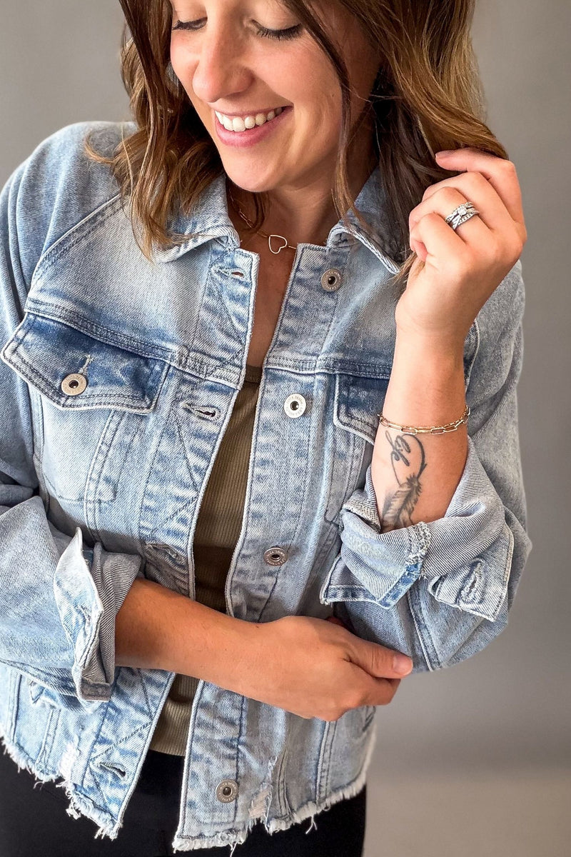 A woman wearing a light-wash blue denim jacket over a dark olive top, shown in a close-up shot against a neutral gray background.