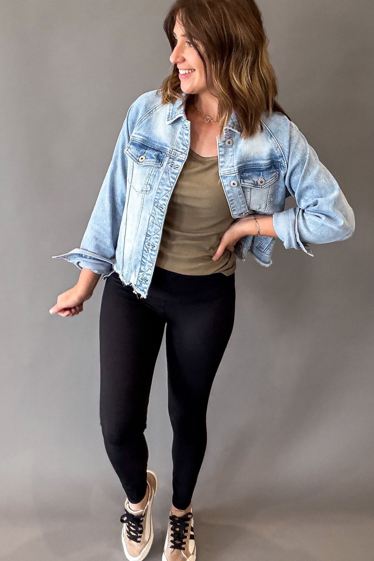 A woman modeling a light-wash blue cropped denim jacket over an olive green tank top, black leggings, and beige sneakers, standing against a solid gray studio background.