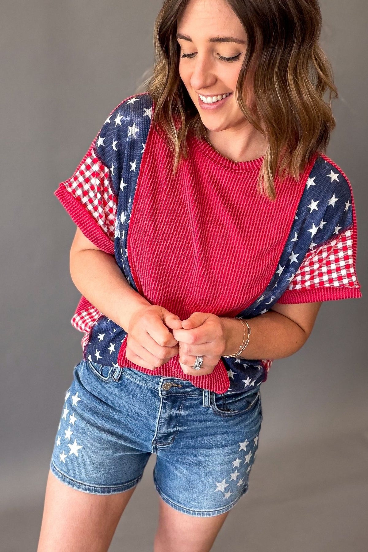 woman wearing a ribbed red horizontal color block short sleeve with blue waffle star print and red gingham print on the sleeves pair with judy blue star shorts