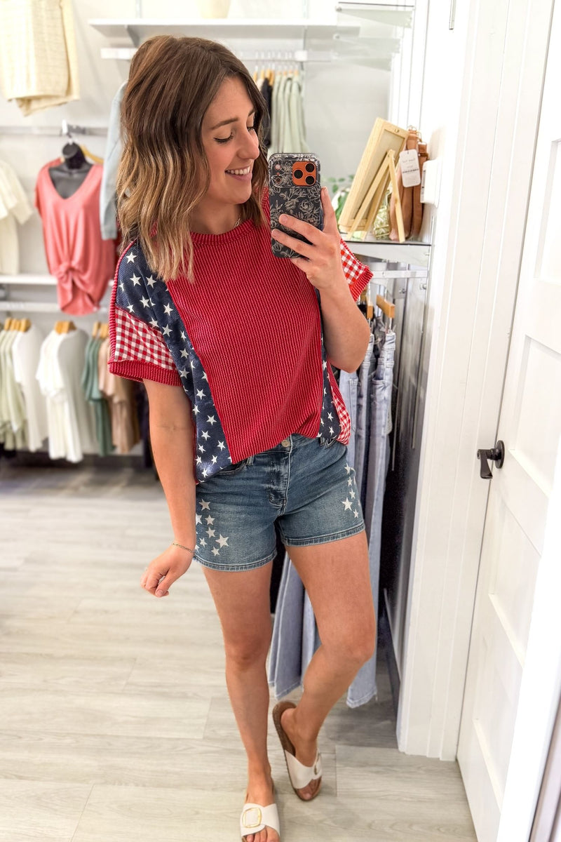 woman taking a mirror selfie wearing a casual relaxed fit, red white and blue ribbed short sleeve with blue jeans and white corkys sandals
