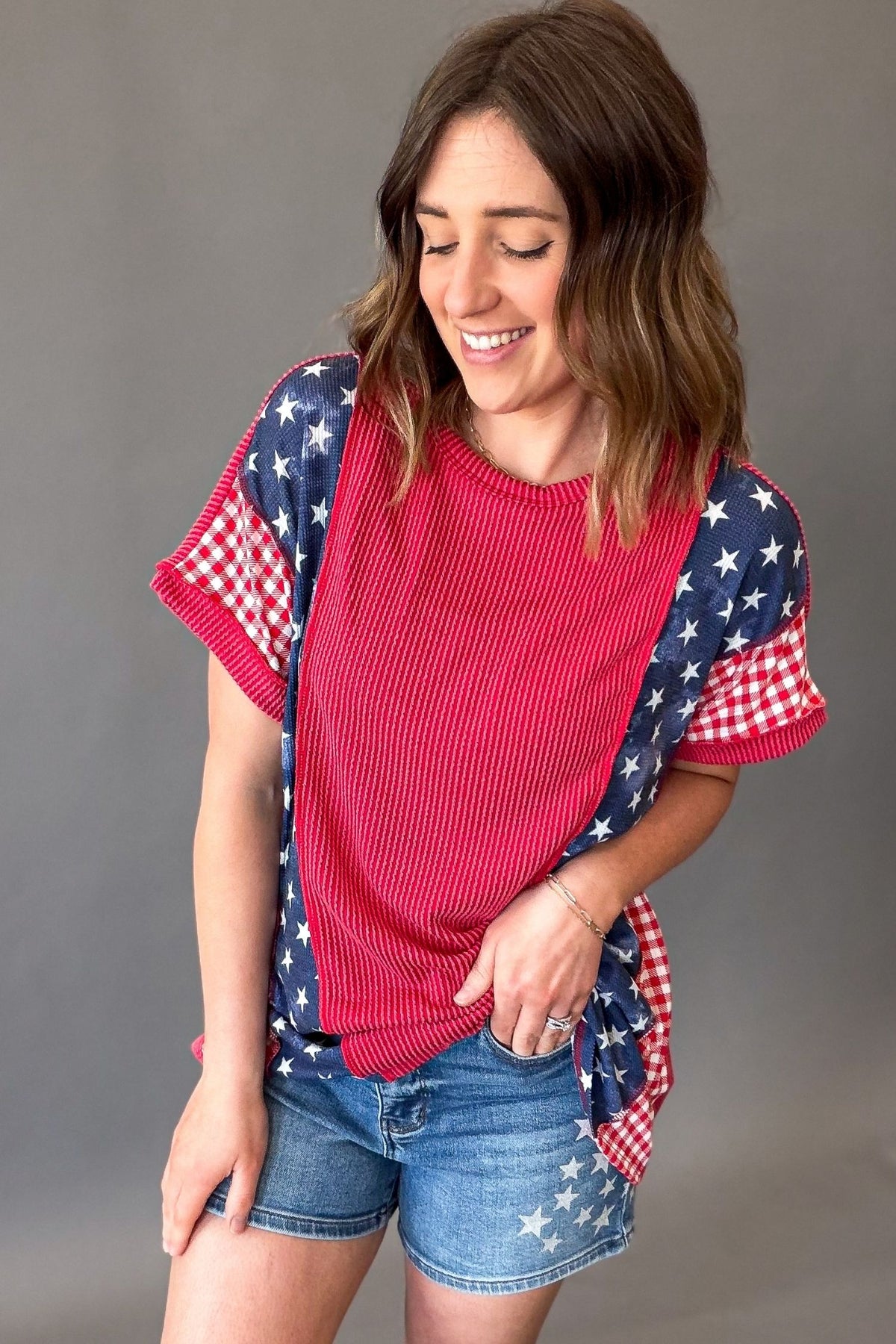 woman wearing a ribbed red horizontal color block short sleeve with blue waffle star print and red gingham print on the sleeves pair with judy blue star shorts untucked to show top length.