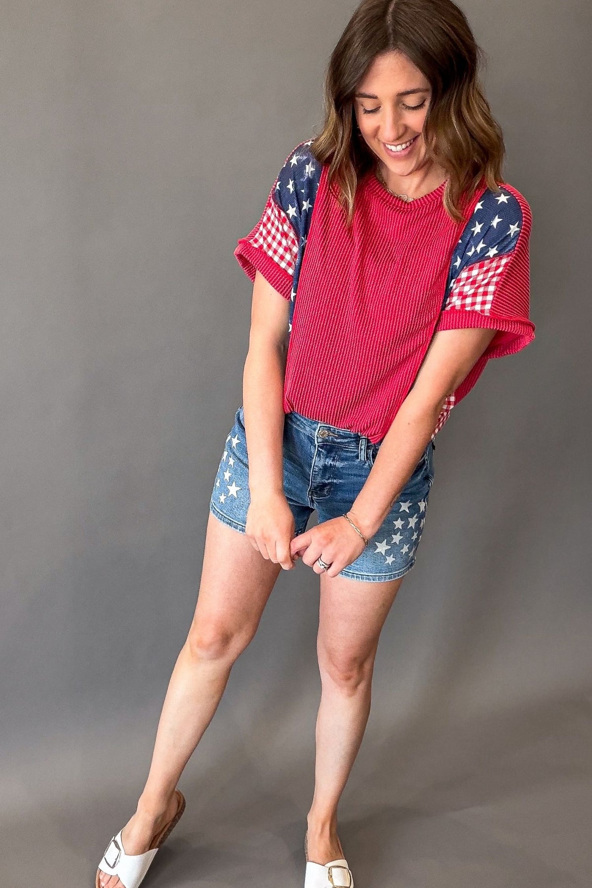 woman wearing a ribbed red horizontal color block short sleeve with blue waffle star print and red gingham print on the sleeves pair with judy blue star shorts