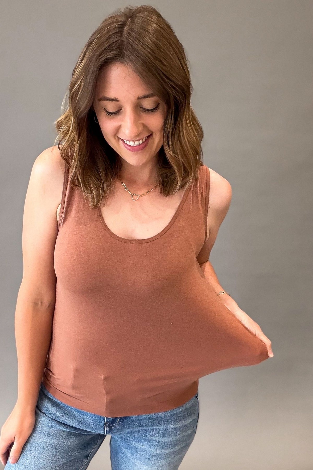 A woman wearing a scoop-neck tank top in a muted terracotta or rust color, paired with blue jeans, posing to show the fabric's stretch against a plain gray studio background.