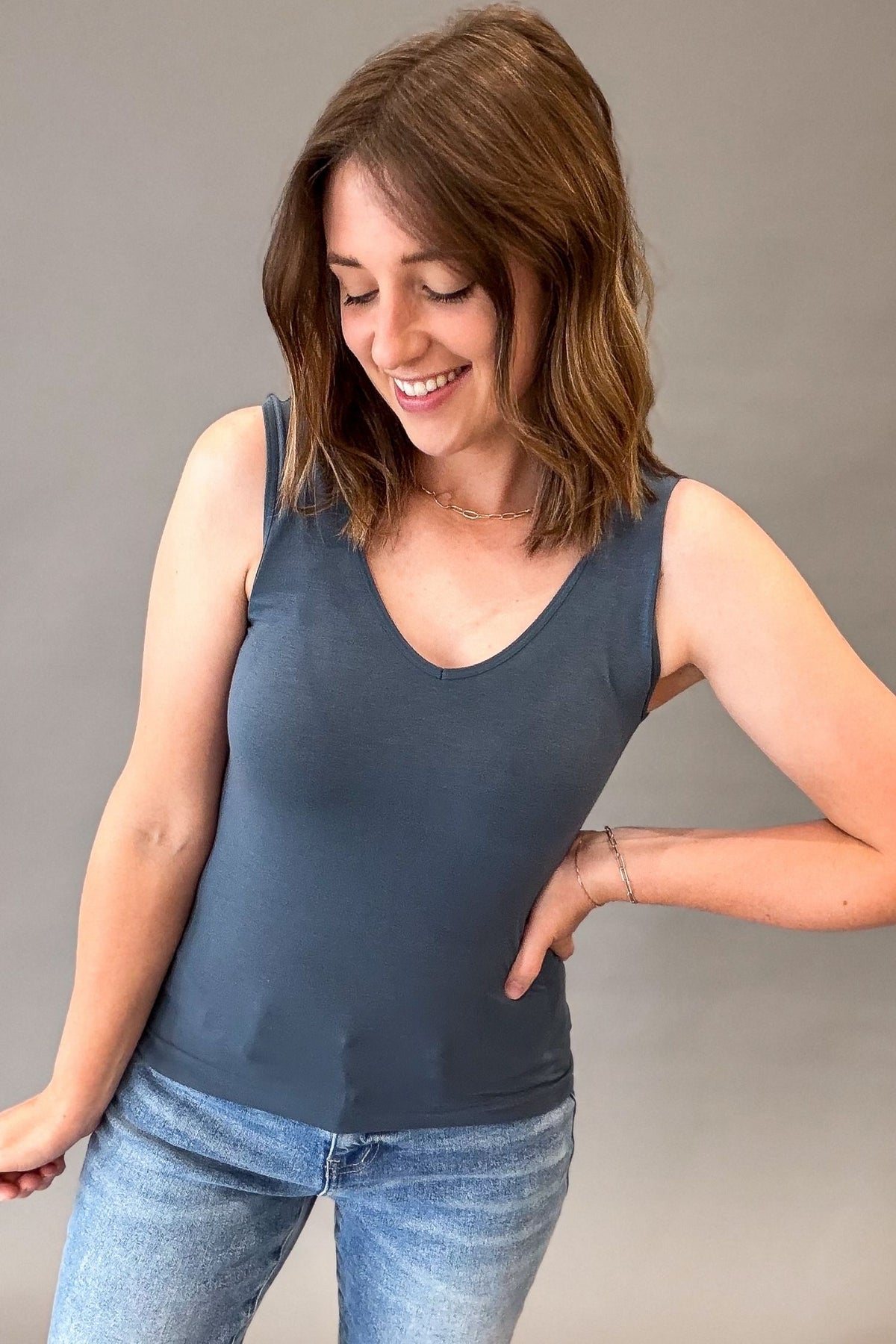 A woman wearing a slate blue-gray sleeveless scoop-neck tank top and light-wash blue jeans, standing against a neutral gray studio background.