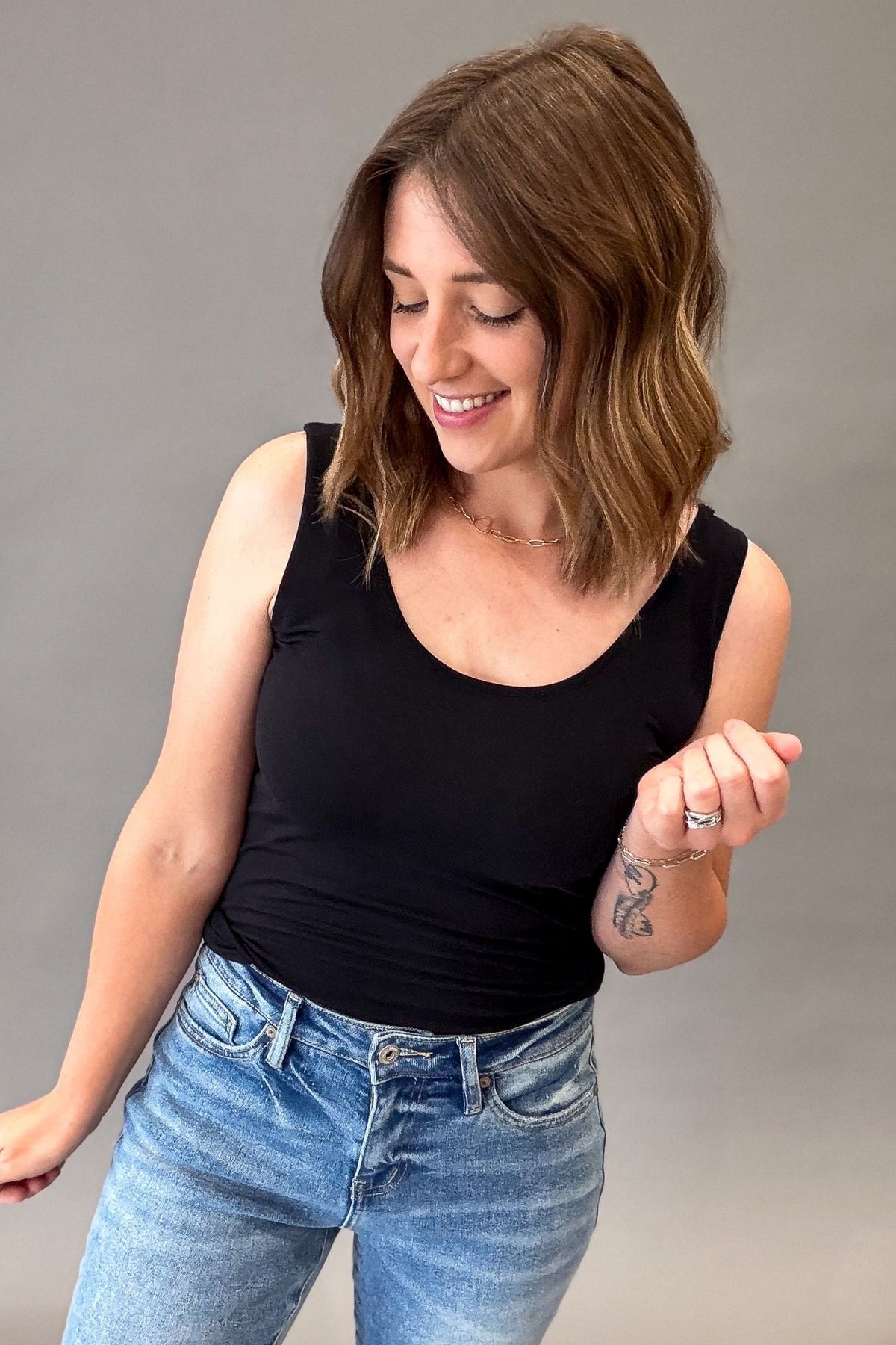 A woman with shoulder-length brown hair is wearing a black scoop-neck tank top and light-wash blue jeans, posing against a solid gray studio background.