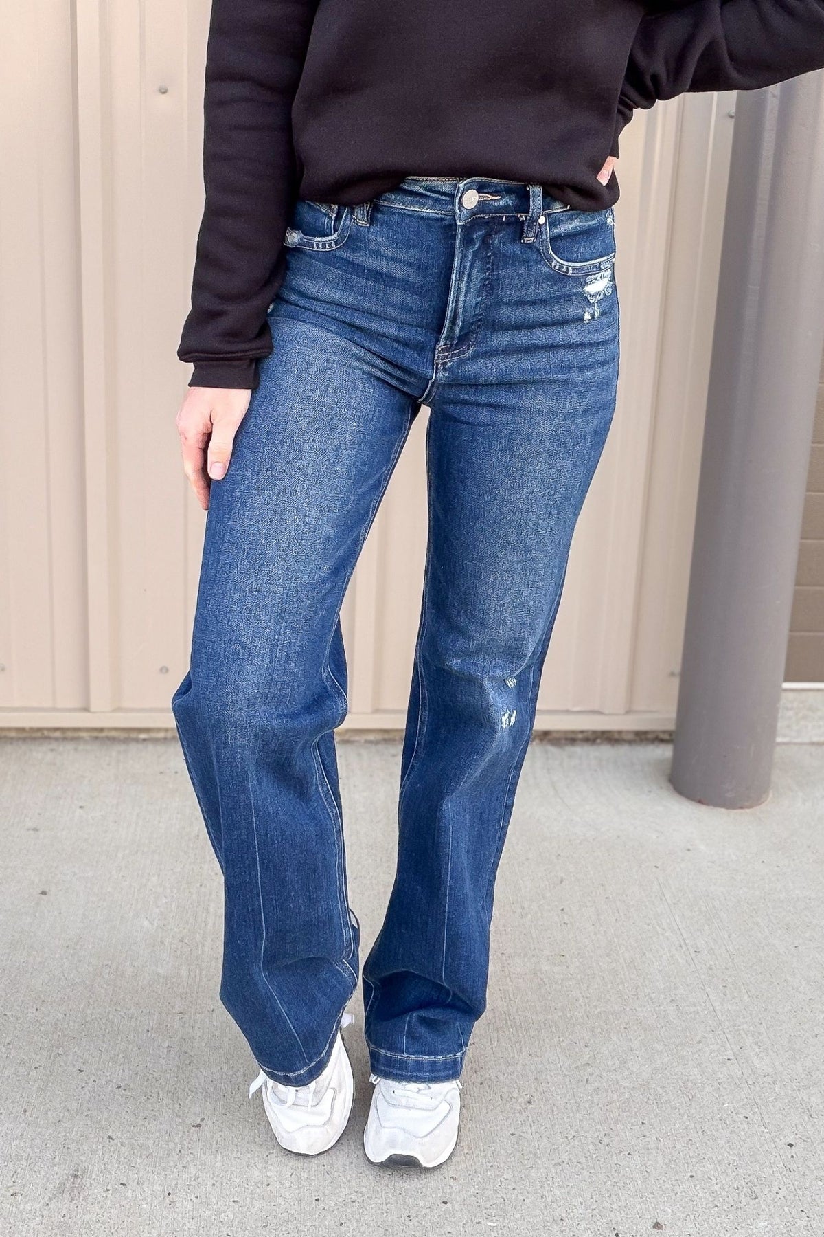 Person wearing blue jeans and a dark jacket standing against a plain background