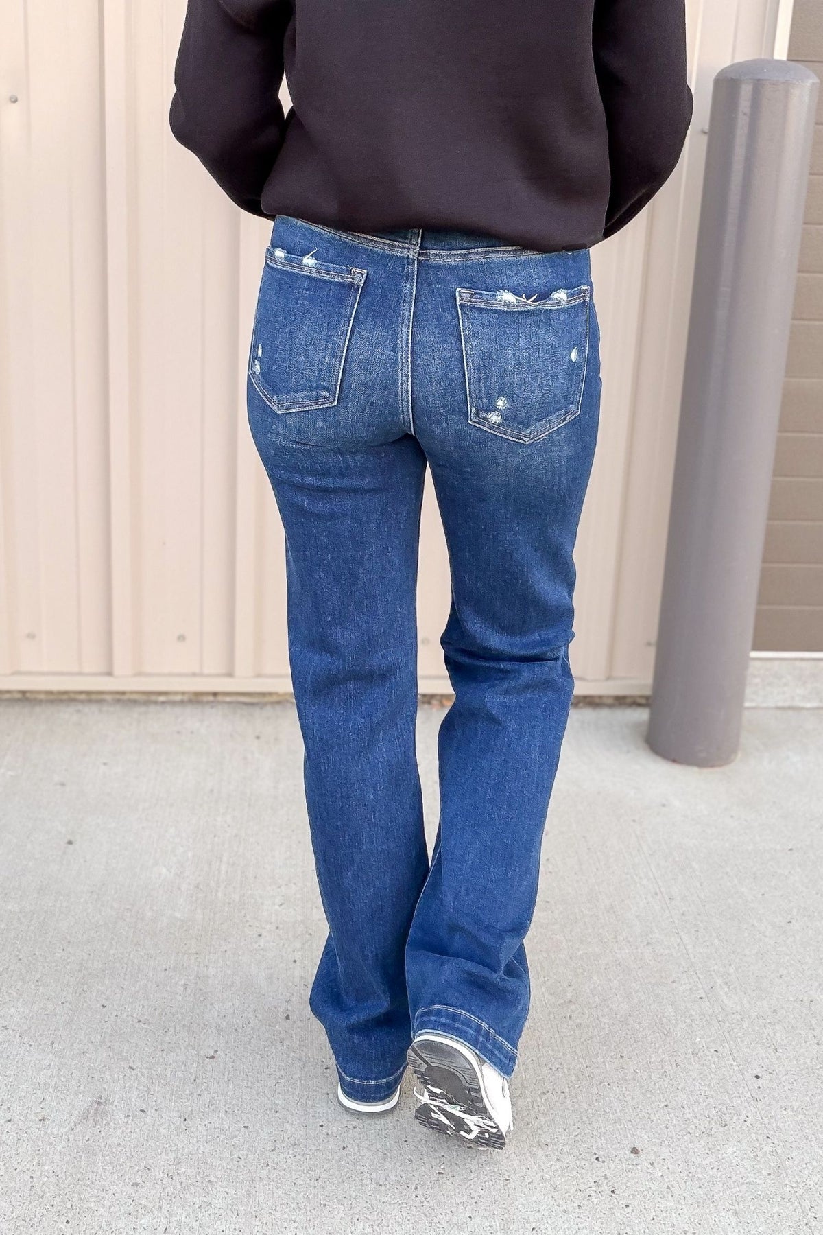 Person wearing blue jeans and a black top standing on a concrete surface with a beige wall in the background.