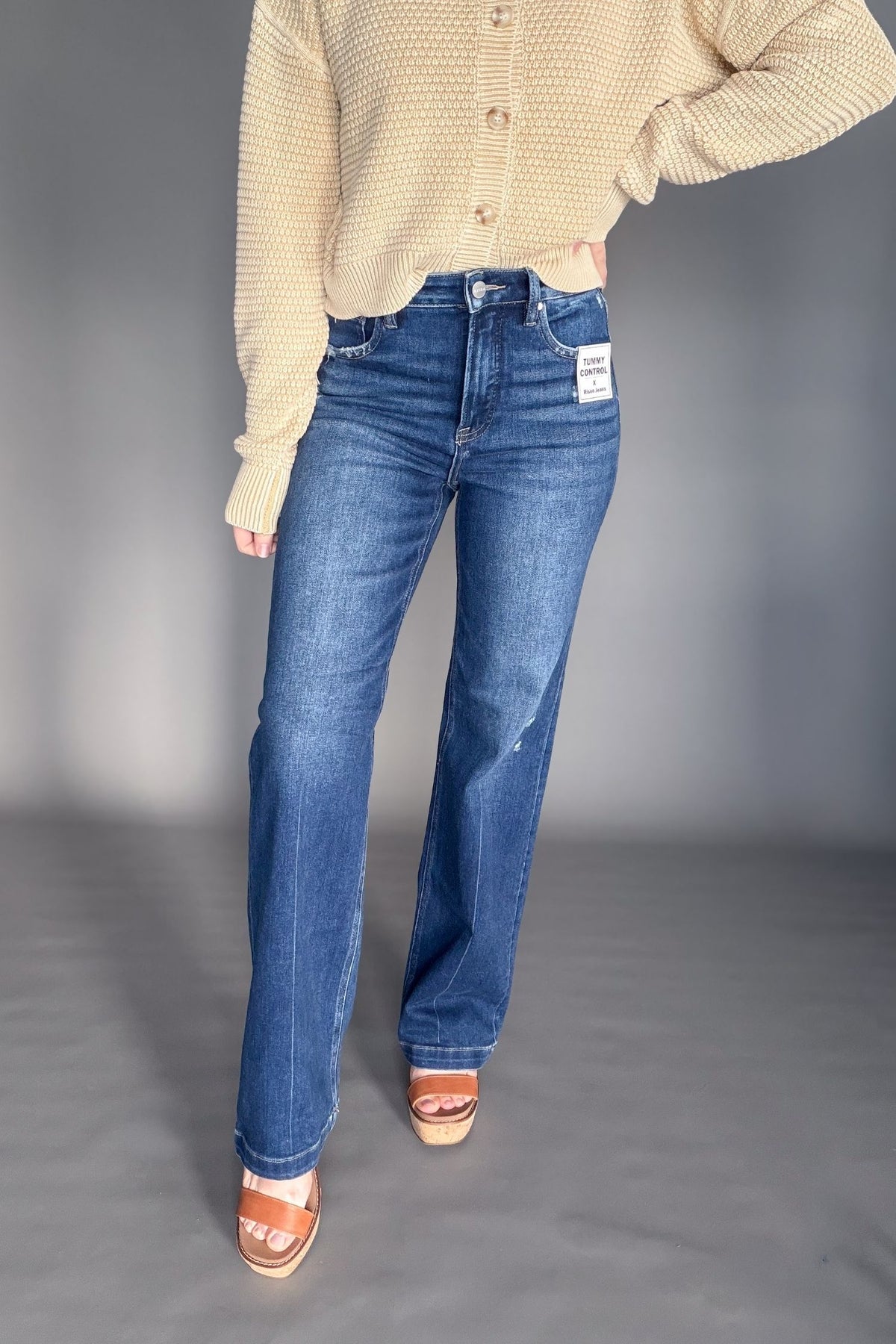 Person wearing blue jeans and a camel cardigan on a gray background