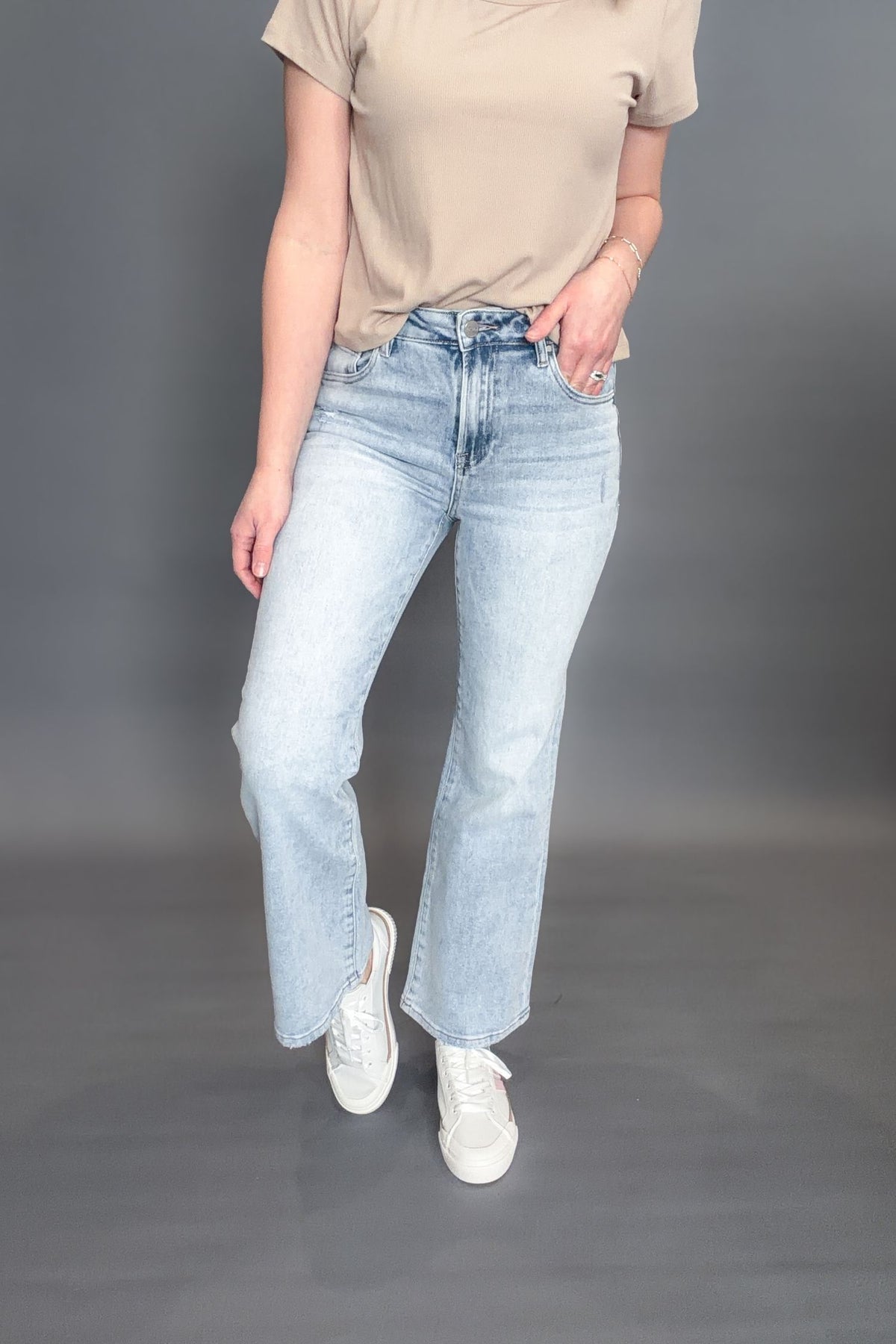 Woman wearing Risen Kathleen High Rise Ankle Flare Jeans on a grey background with white sneakers and a taupe tee showing the front view of the jeans.