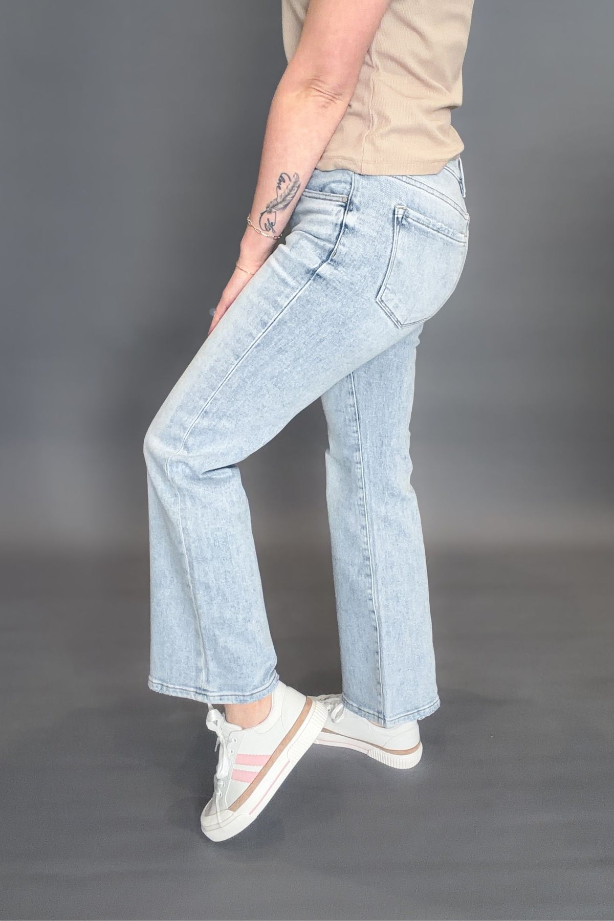 Woman wearing Risen Kathleen High Rise Ankle Flare Jeans on a grey background with white sneakers and a taupe tee showing the side view of the jeans