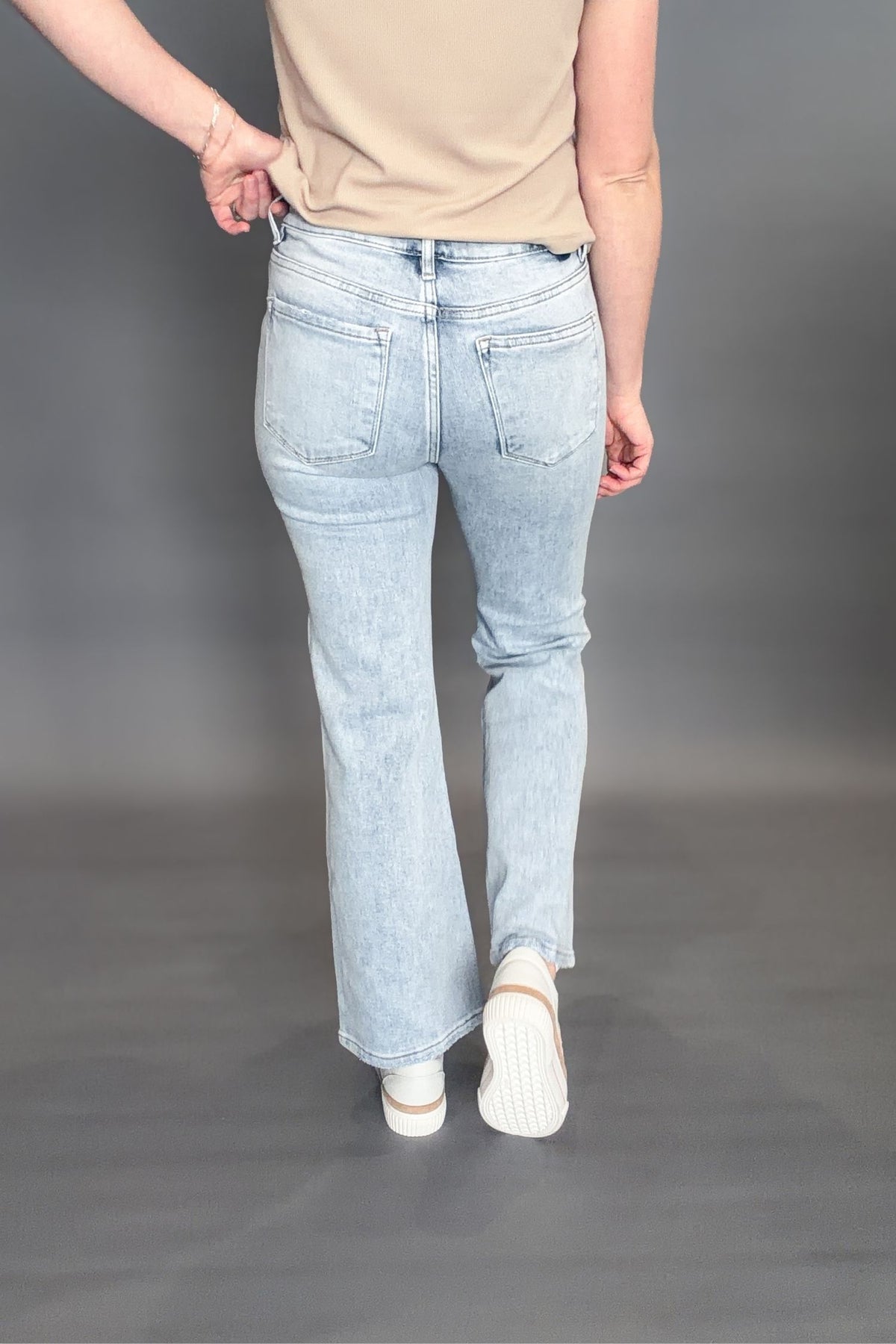 Woman wearing Risen Kathleen High Rise Ankle Flare Jeans on a grey background with white sneakers and a taupe tee showing the back view of the jeans