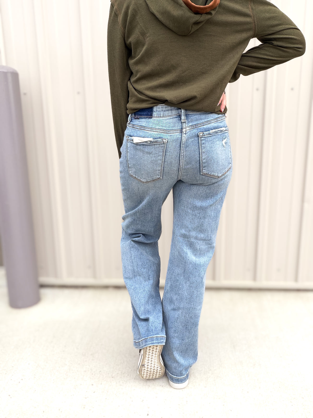 Person wearing a green hoodie and blue jeans standing against a white wall.