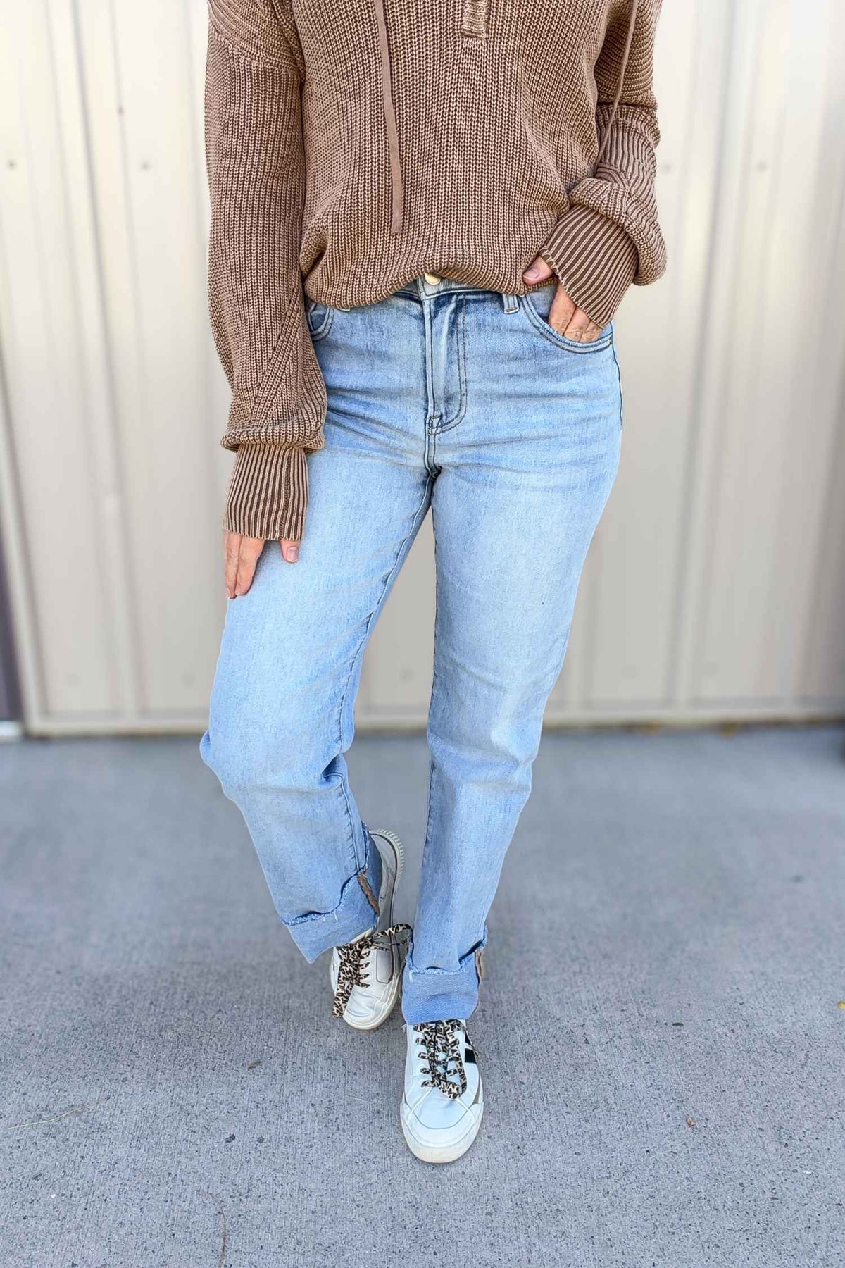 Person wearing a brown sweater, light blue jeans, and white sneakers against a plain background