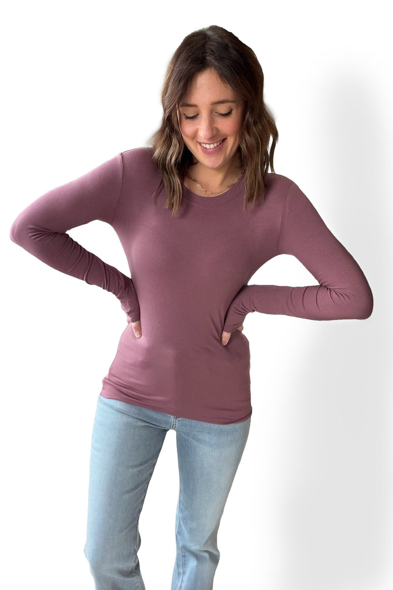 Woman wearing a mauve/rose colored ribbed long-sleeve top and light blue jeans on a white background