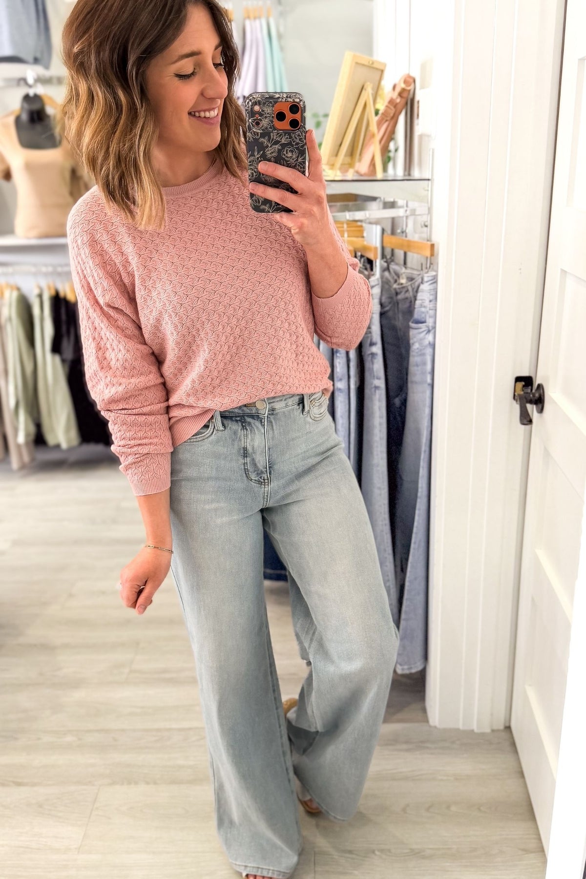 Woman taking a mirror selfie wearing a pink sweater and light blue jeans in a clothing store.