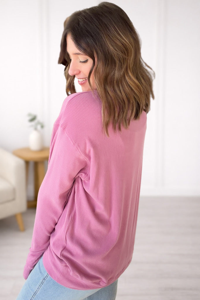 Woman wearing a pink long-sleeve top in a room with a white wall and a small table.