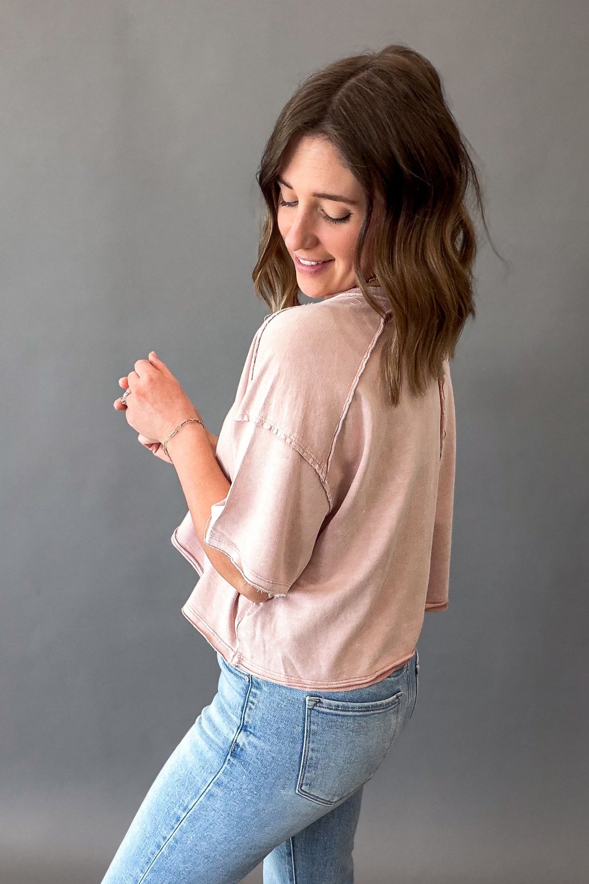 woman wearing a light pink short sleeve cropped tee with blue jeans on a grey background