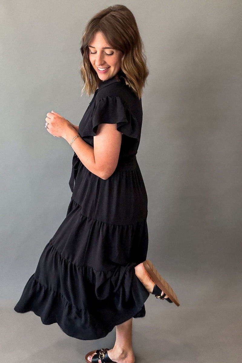 A woman wearing a black tiered midi dress with short flutter sleeves, shown from a side-back angle against a plain gray studio background.