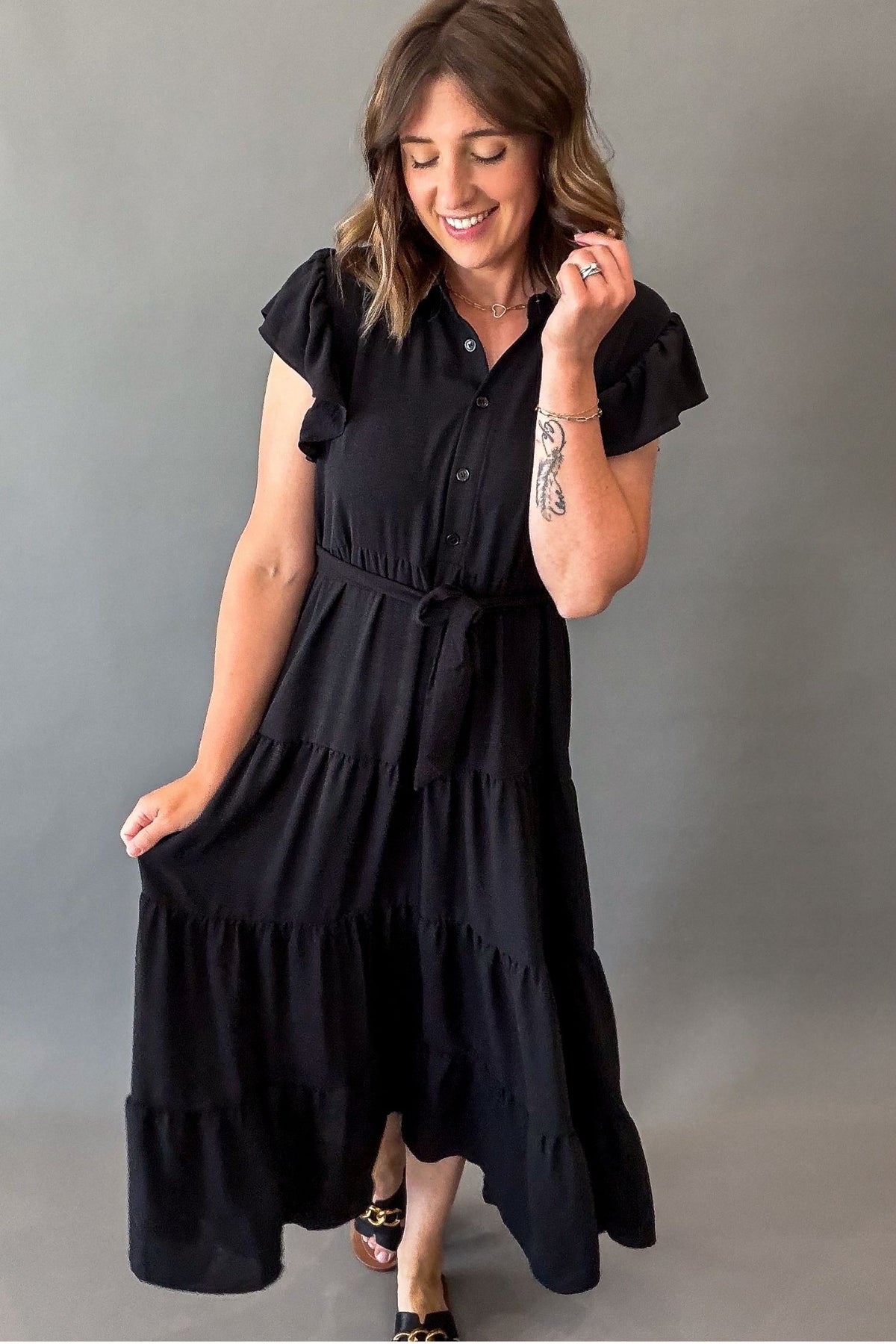 A woman modeling a black tiered midi dress with ruffled short sleeves, a buttoned collar, and a tie waist, photographed against a plain gray studio background.