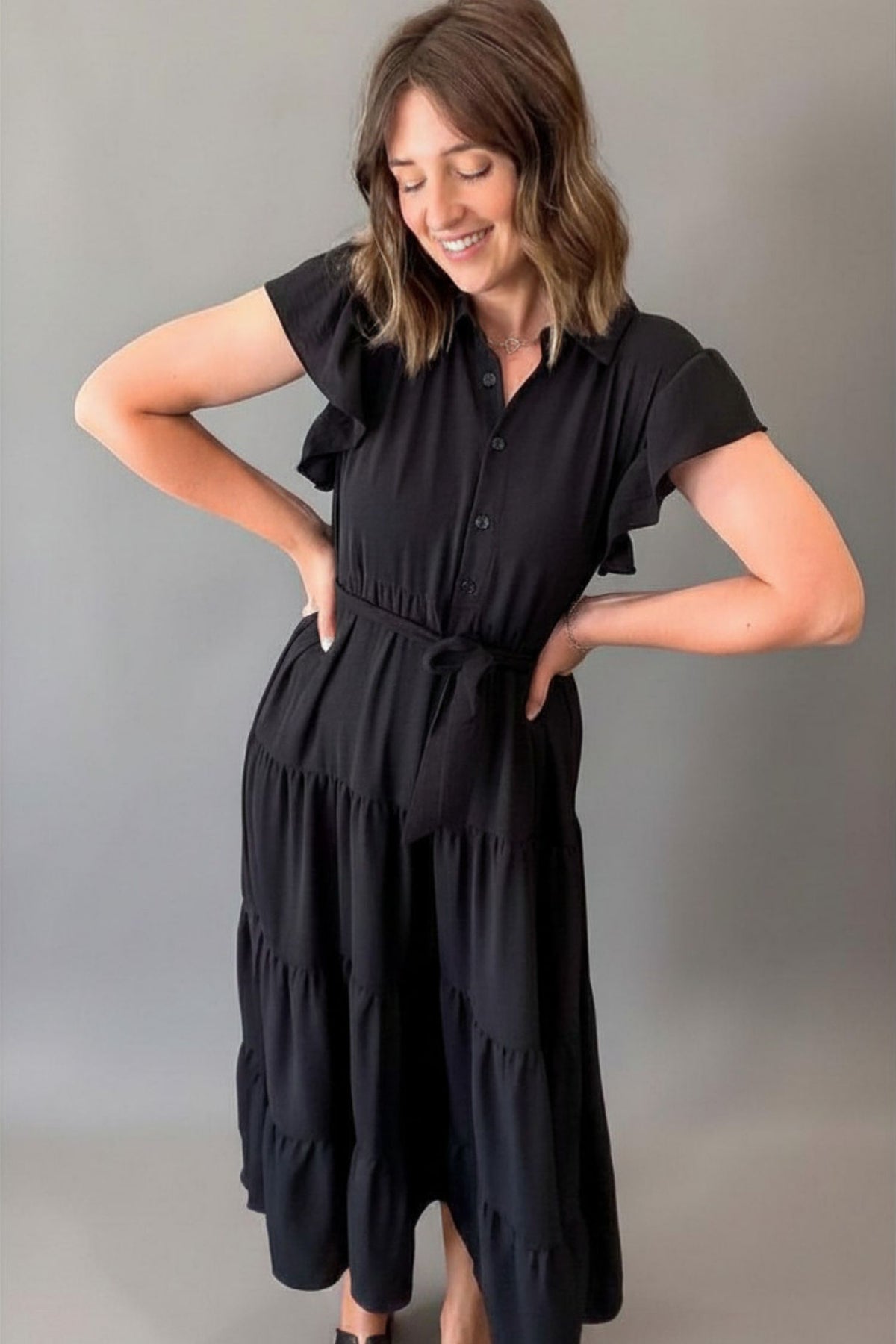 Woman wearing a flowy black button-front dress with ruffled sleeves, a tie waist, and a tiered skirt styled for an effortlessly elegant look.
