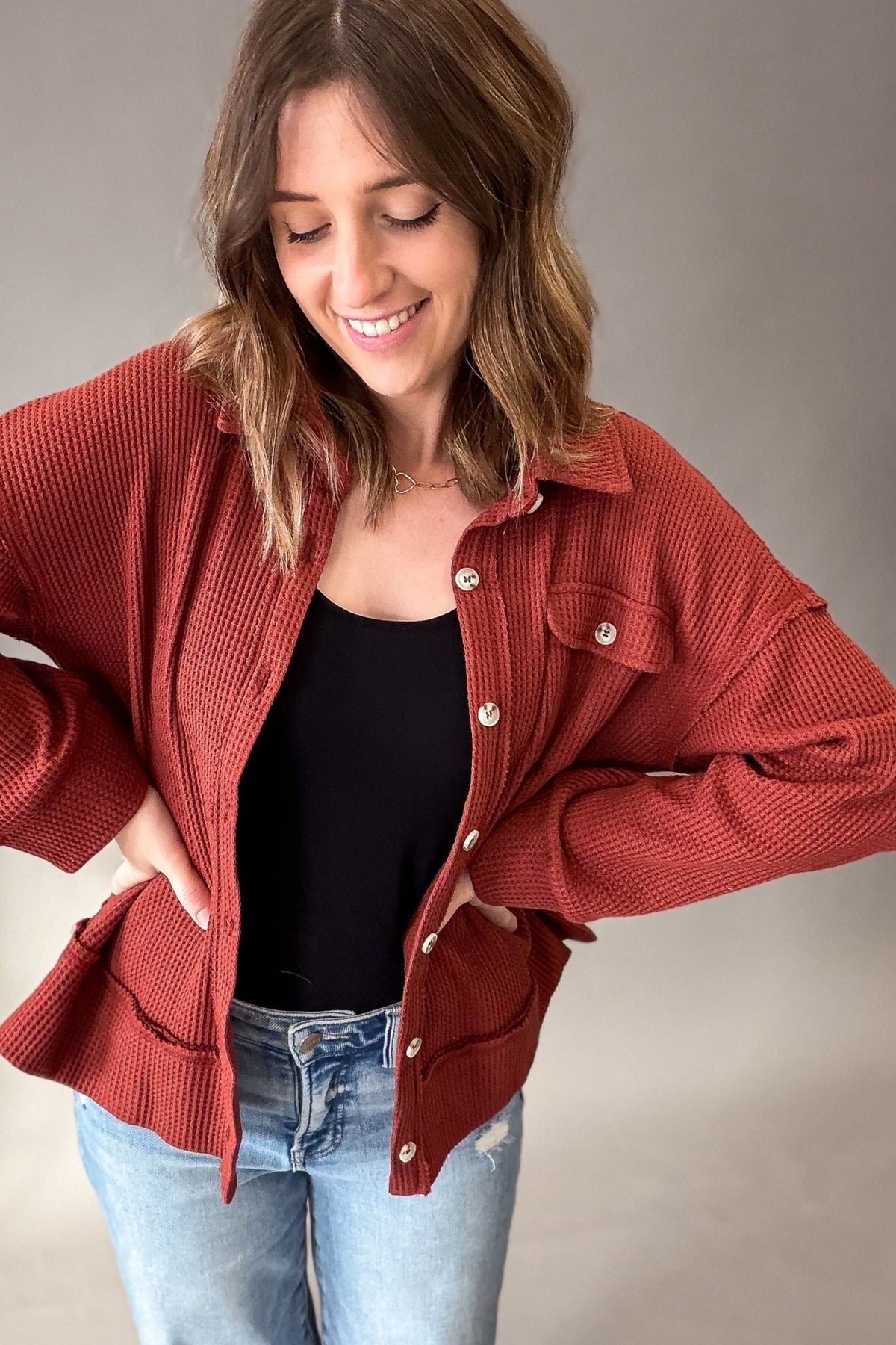 A woman wearing a rust-red waffle-knit button-up cardigan over a black top and light-wash blue jeans, posing against a neutral gray studio background.