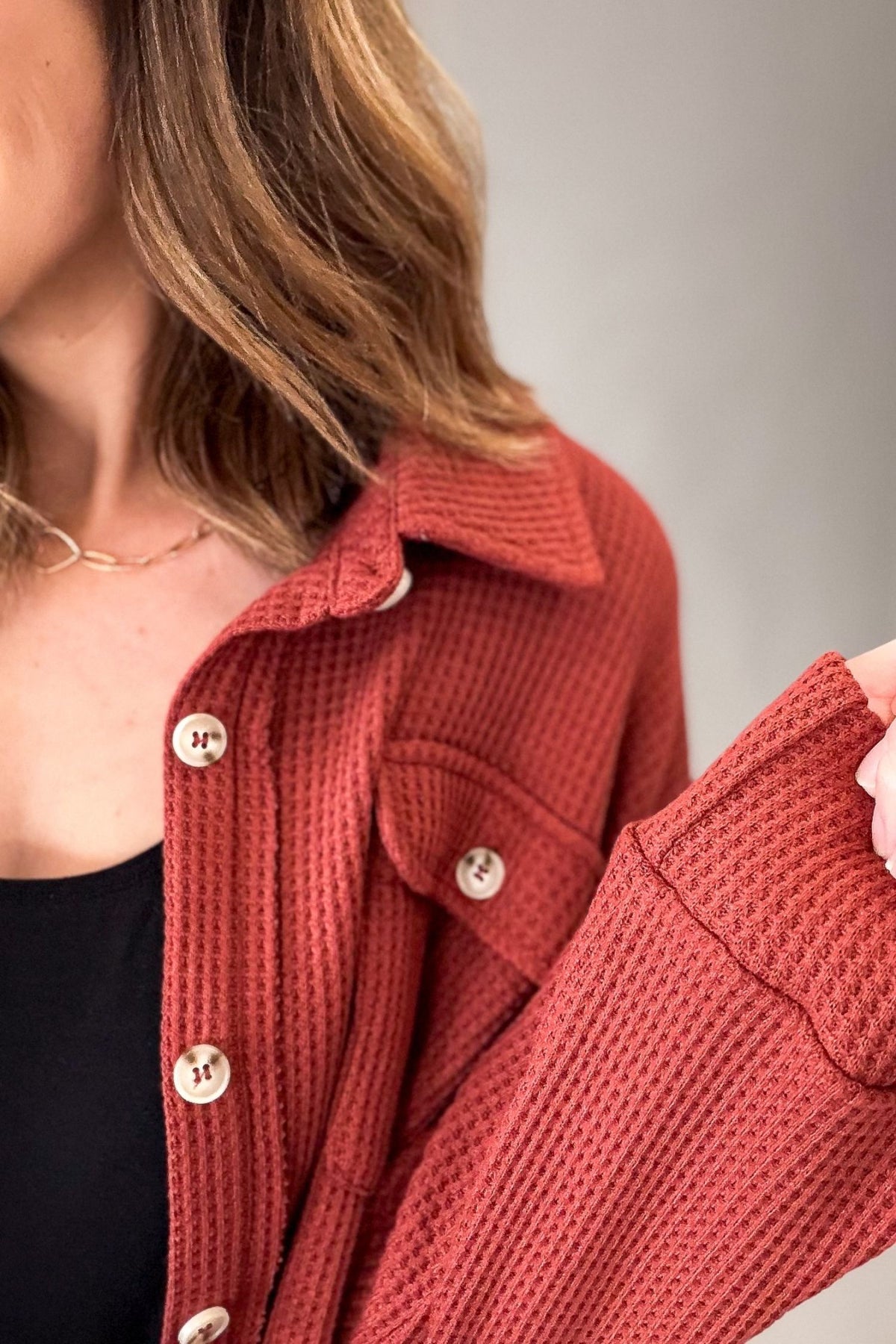 A close-up of a woman wearing a textured waffle-knit cardigan in a muted terracotta or rust red color, paired with a black top and a delicate gold necklace.