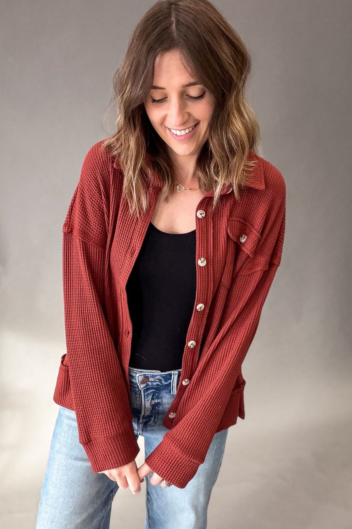 A woman wearing a rust red textured button-up shirt over a black top and light blue jeans, standing against a neutral gray studio background.