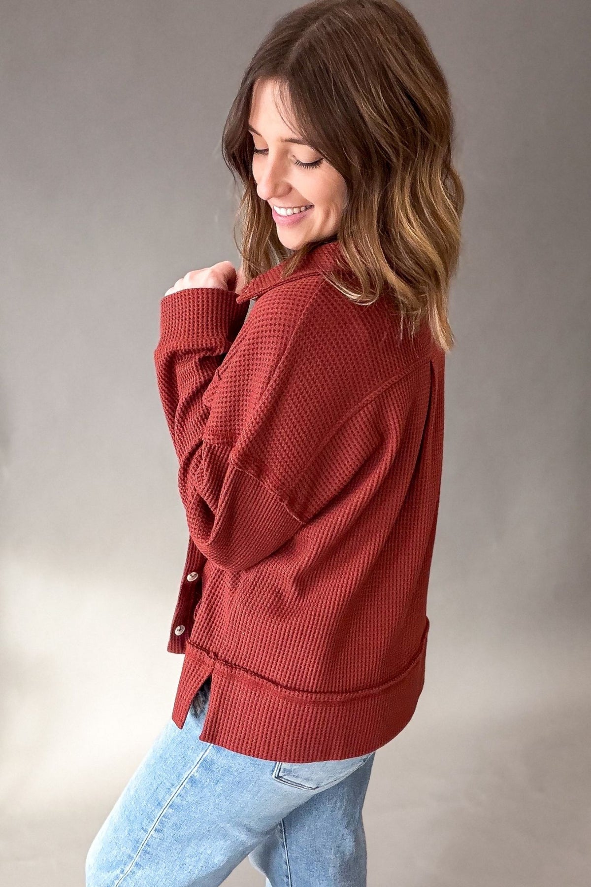 A woman wearing a rust-red waffle-knit button-up shirt and light blue jeans, photographed against a neutral gray studio background.