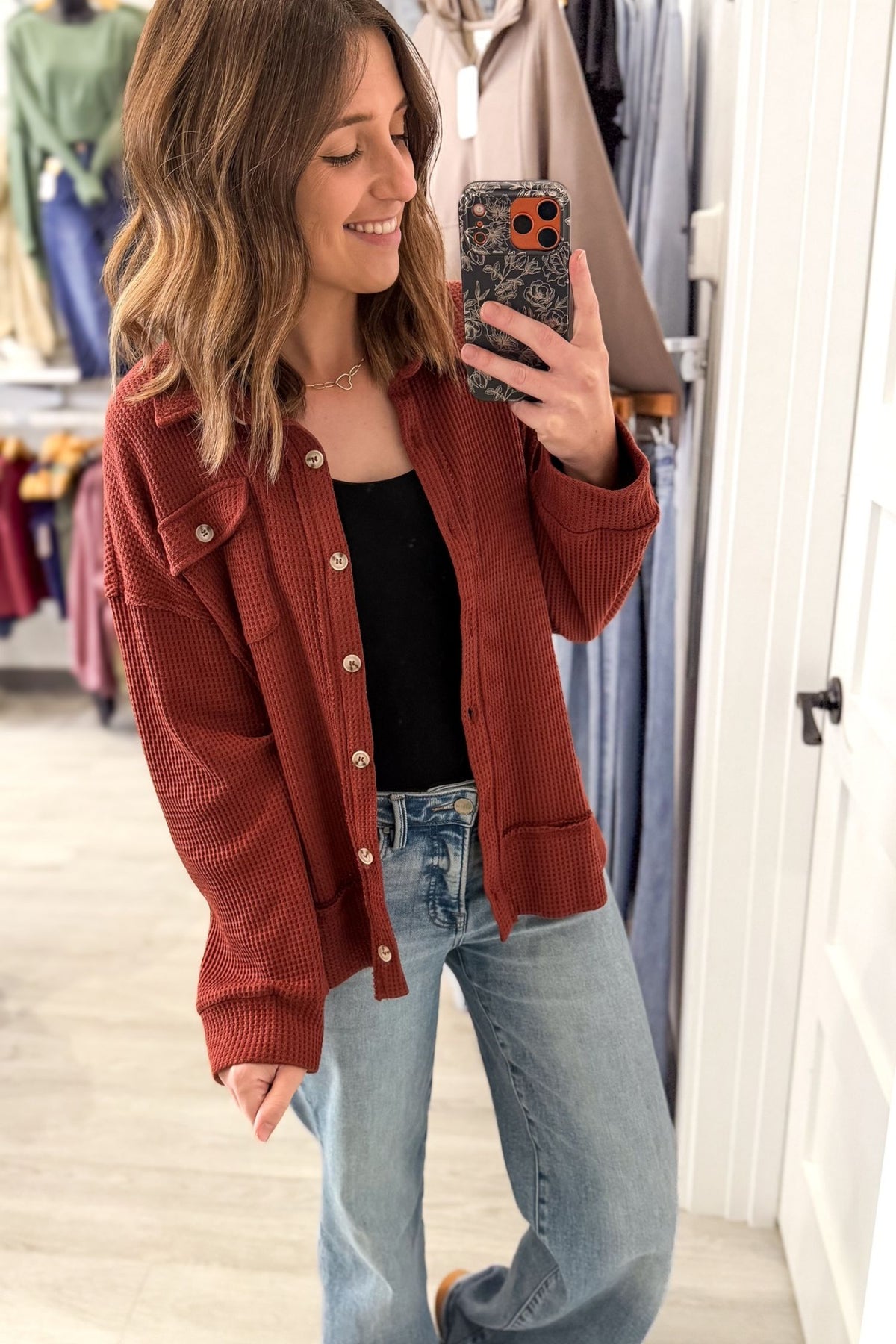 A woman takes a mirror selfie in a clothing store, wearing a rust-colored waffle-knit cardigan over a black top and light-wash blue jeans.