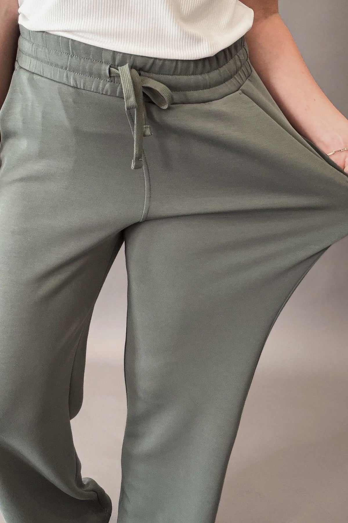 woman wearing a sage lounge pant paired with a white shirt and neutral colored sneakers against a charcoal backdrop showing the stretch in the pant