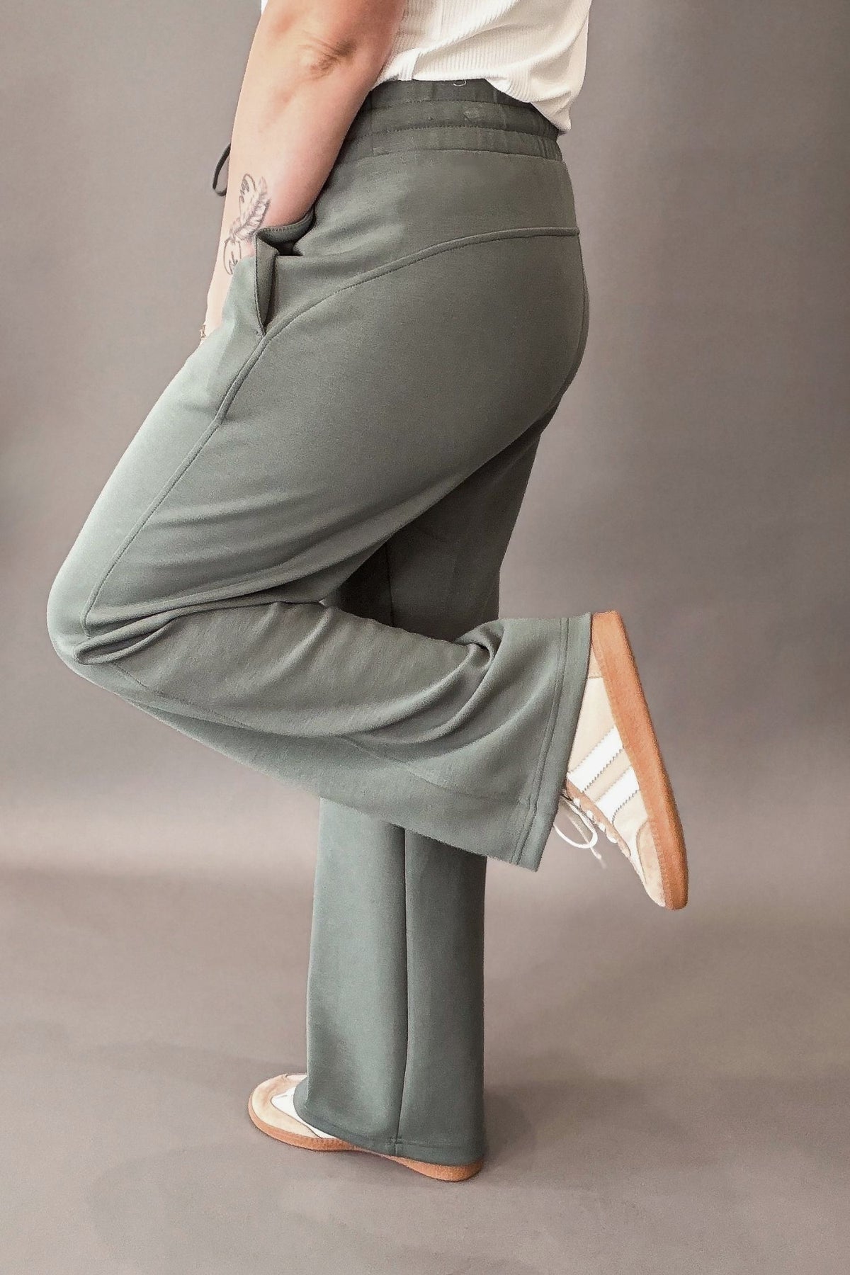 woman wearing a sage lounge pant paired with a white shirt and neutral colored sneakers against a charcoal backdrop showing the side details of the pant.