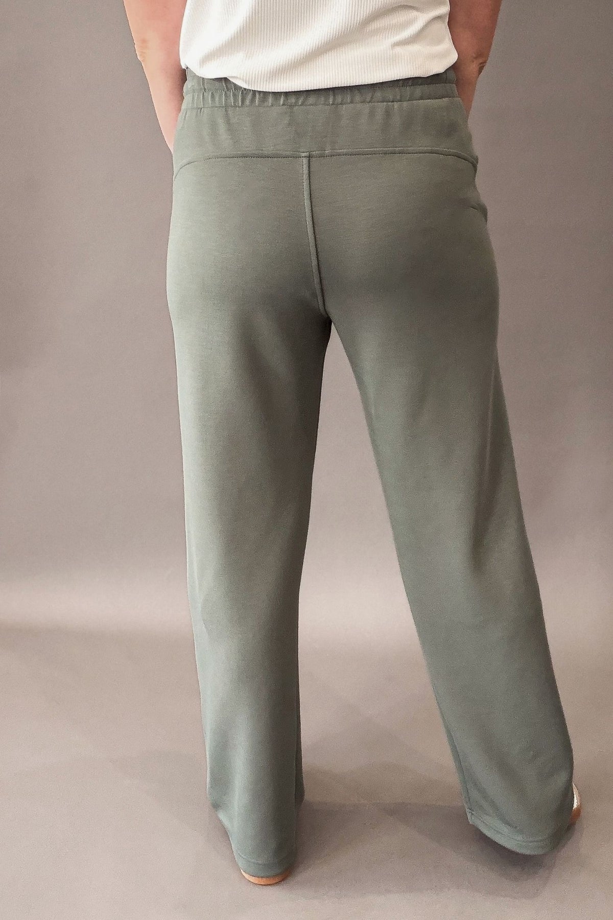 woman wearing a sage lounge pant paired with a white shirt and neutral colored sneakers against a charcoal backdrop showing the back details of the pant