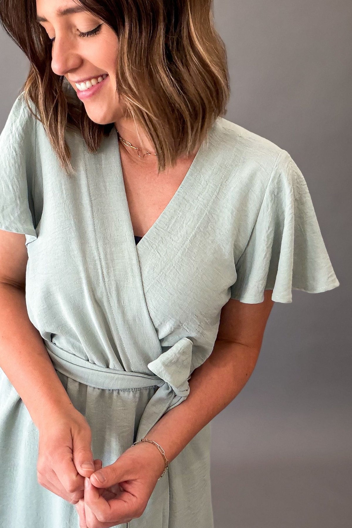 Woman wearing a soft sage green midi dress featuring flutter sleeves, a faux wrap neckline, adjustable tie waist, side pockets, and a flowy lined skirt.