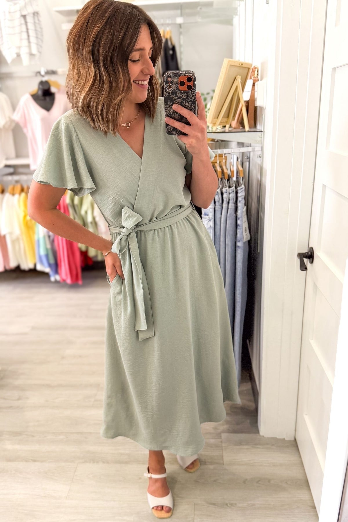 Woman taking a selfie wearing a soft sage green midi dress featuring flutter sleeves, a faux wrap neckline, adjustable tie waist, side pockets, and a flowy lined skirt.
