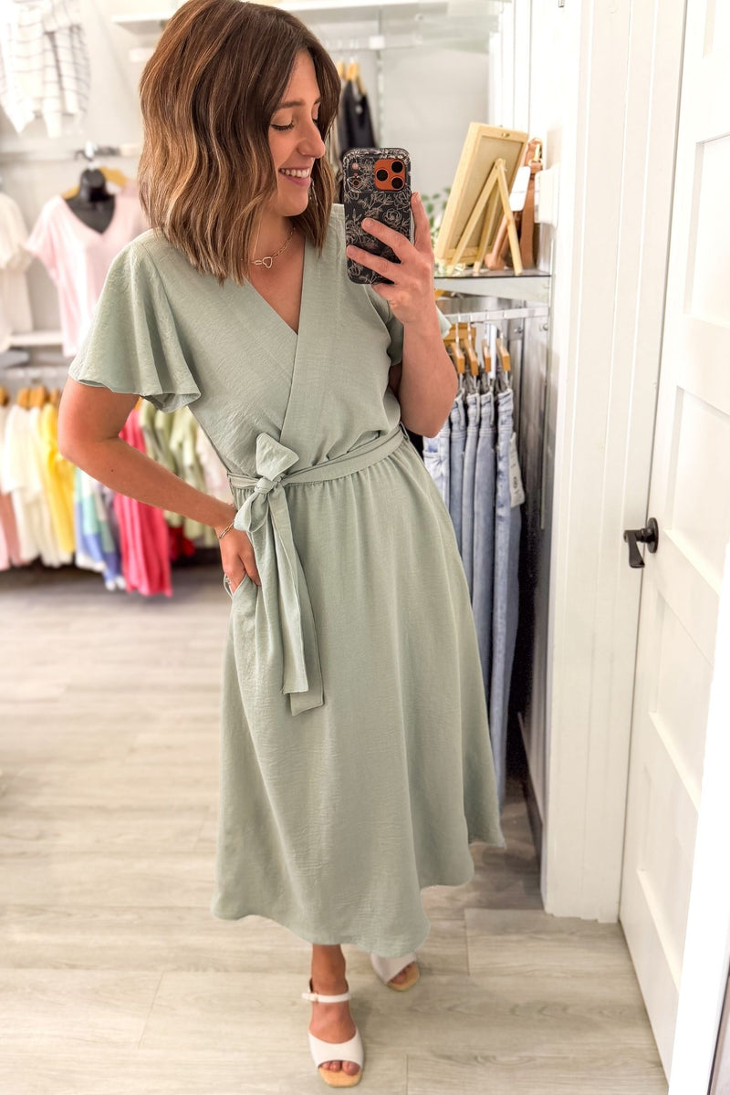 Woman taking a selfie wearing a soft sage green midi dress featuring flutter sleeves, a faux wrap neckline, adjustable tie waist, side pockets, and a flowy lined skirt.