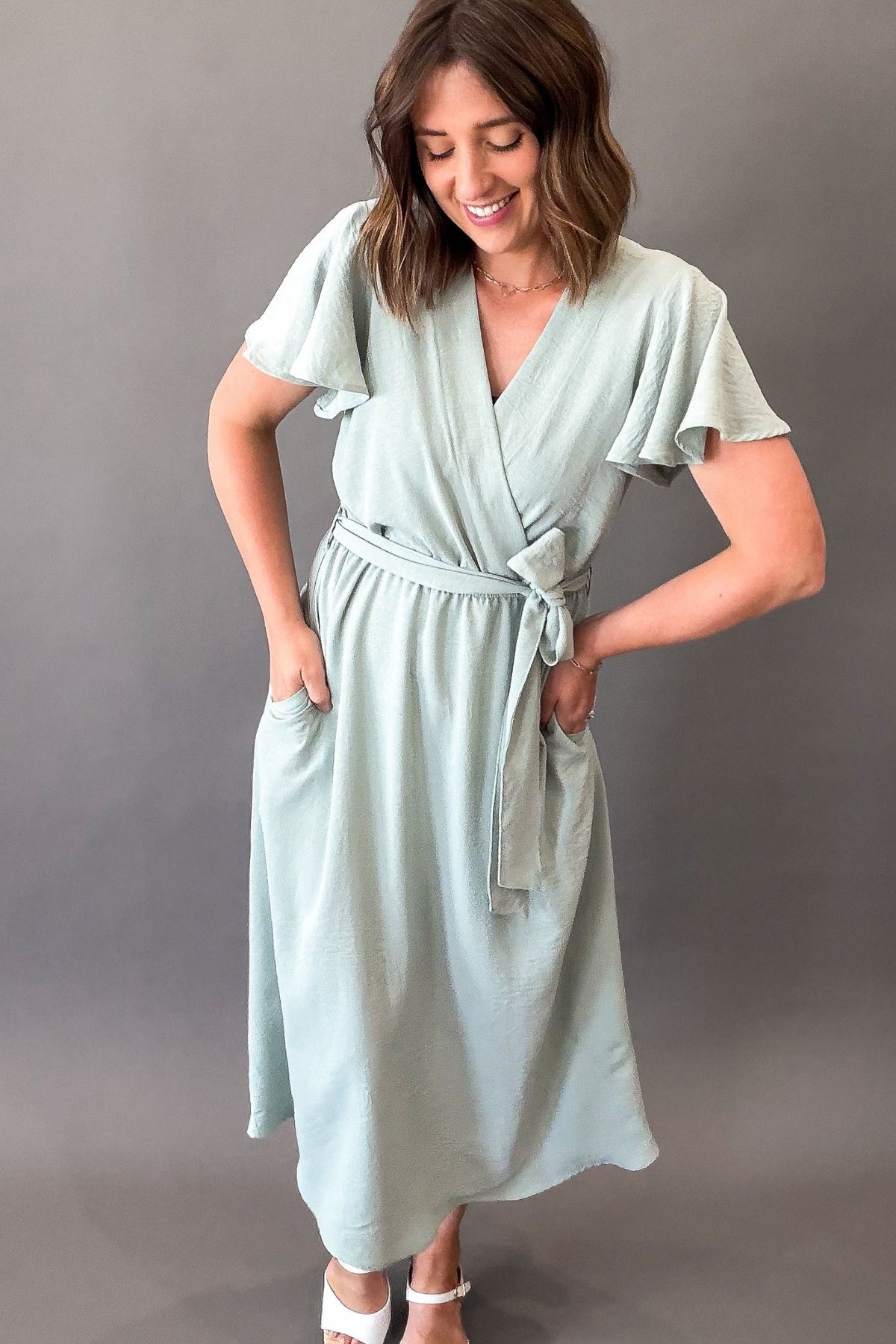 Woman wearing a soft sage green midi dress featuring flutter sleeves, a faux wrap neckline, adjustable tie waist, side pockets, and a flowy lined skirt.