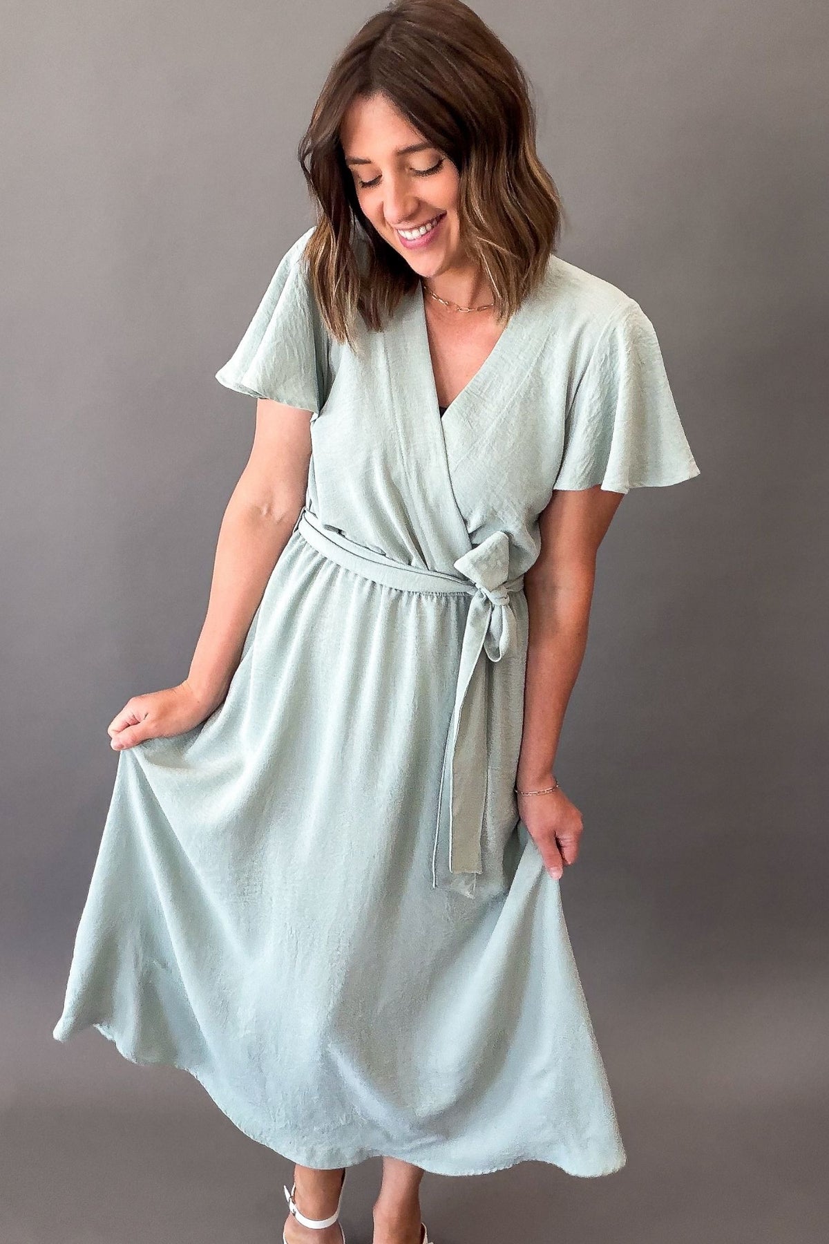 Woman wearing a soft sage green midi dress featuring flutter sleeves, a faux wrap neckline, adjustable tie waist, side pockets, and a flowy lined skirt.