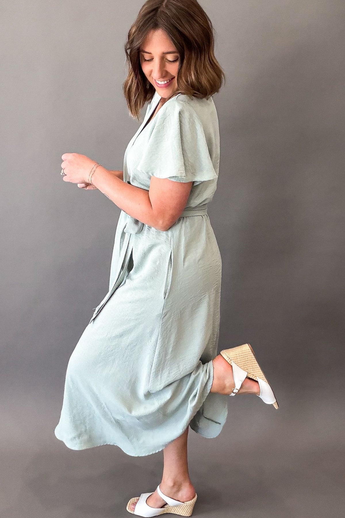 Woman wearing a soft sage green midi dress featuring flutter sleeves, a faux wrap neckline, adjustable tie waist, side pockets, and a flowy lined skirt.