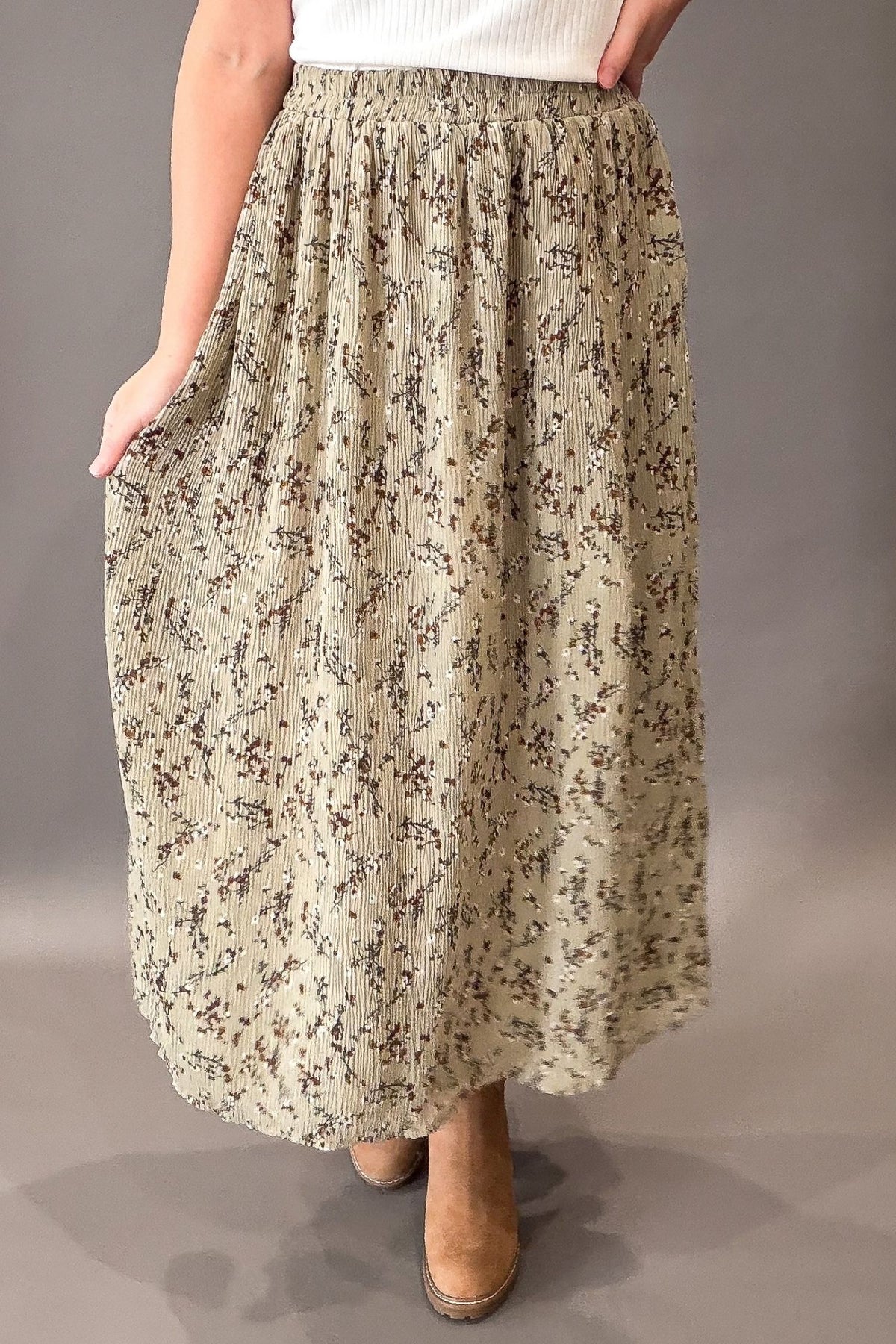 A woman wearing a long, pleated maxi skirt with a beige and brown floral pattern, paired with a white top and tan boots, shown against a neutral gray studio background.