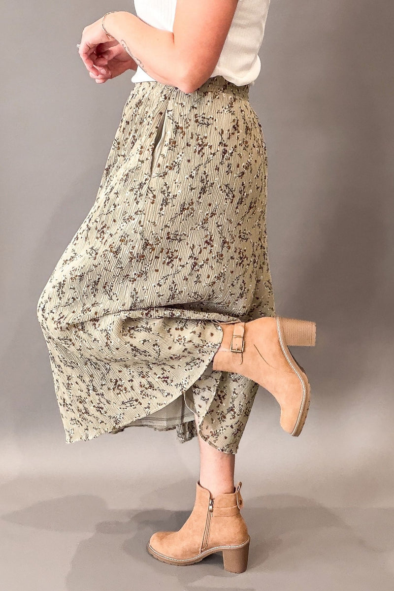 A person wearing a loose, high-waisted midi skirt with a sage and brown floral print, styled with a white top and tan ankle boots against a neutral gray studio background.
