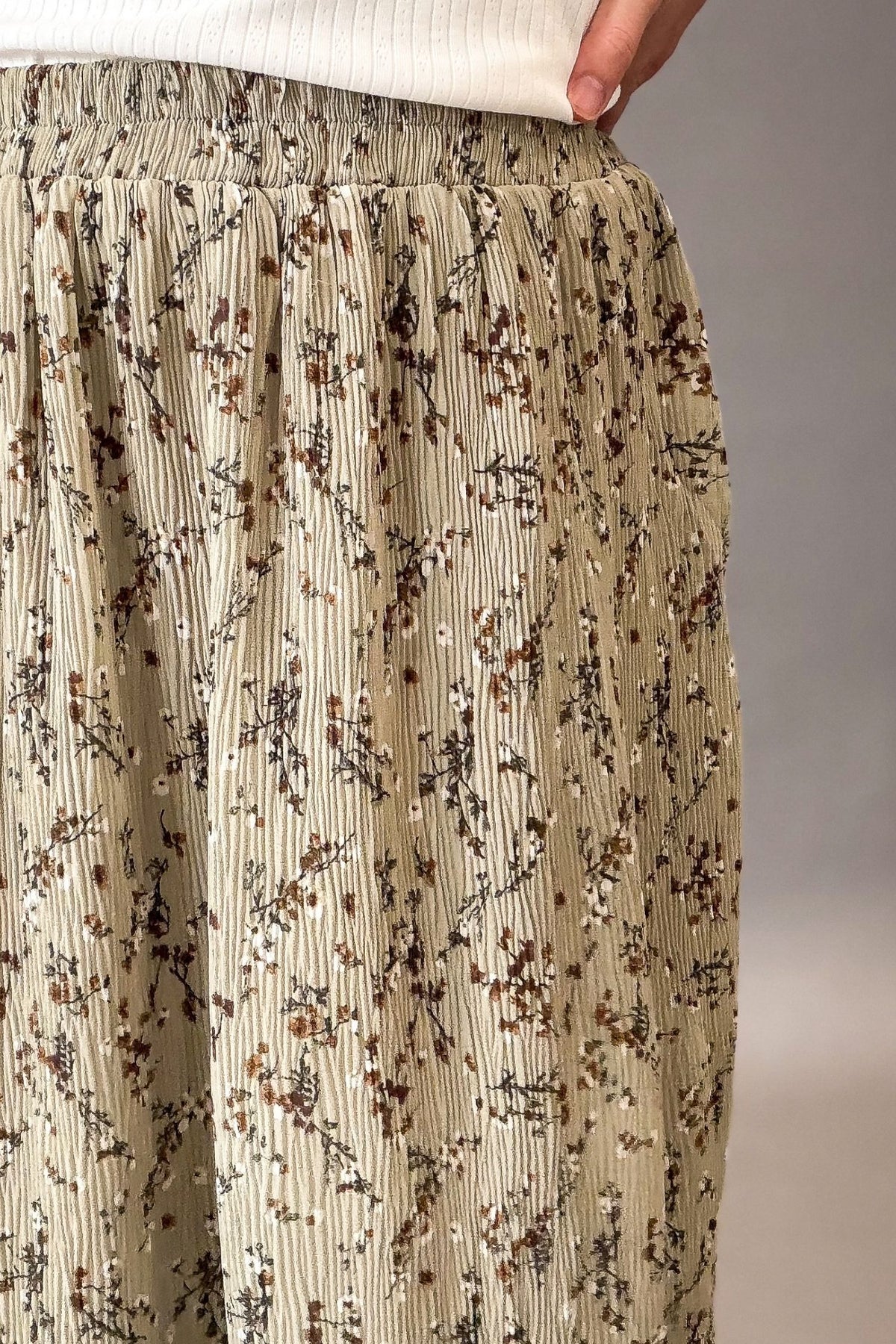 Close-up of a person wearing pleated maxi skirt in a sage green base with a delicate brown, white and black floral branch print, paired with a white ribbed top.