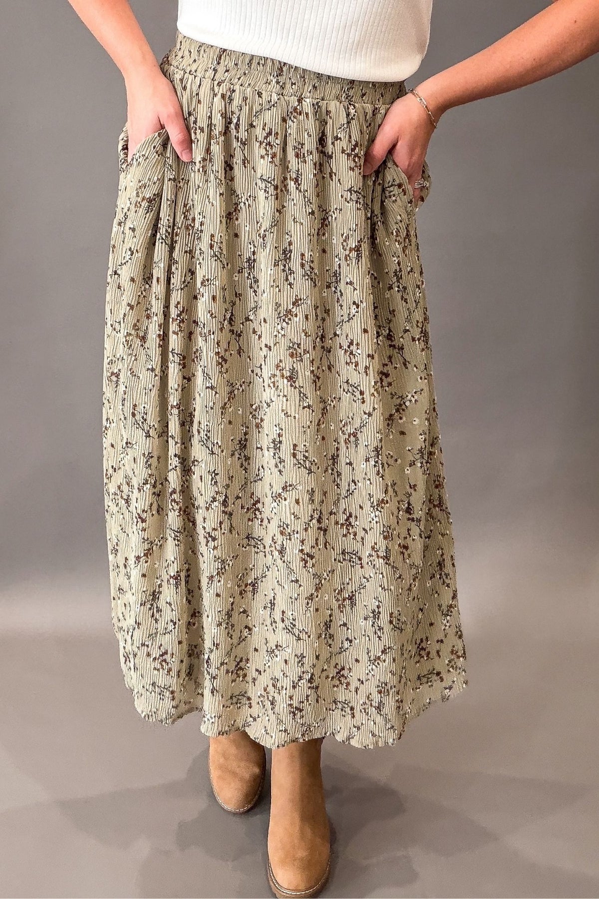 A woman wearing a long, pleated sage maxi skirt with a beige and brown floral pattern, paired with a white ribbed top and tan ankle boots, shown against a neutral gray studio background.
