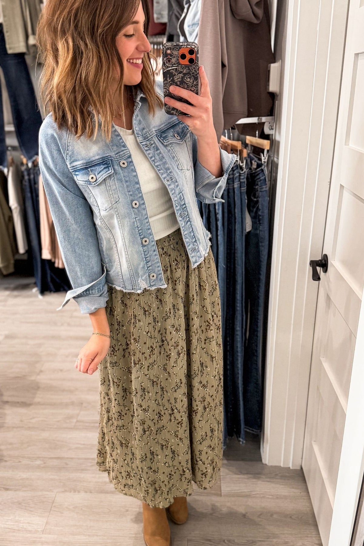 A woman takes a mirror selfie in a clothing boutique wearing a light-wash blue denim jacket over a white top and a long olive green floral midi skirt, paired with tan boots.