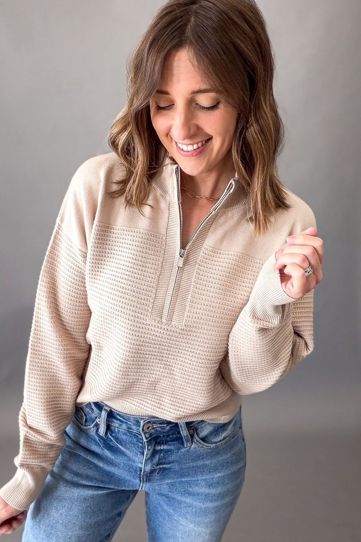 A woman wearing a beige textured quarter-zip pullover sweater and blue jeans, smiling and looking down against a plain gray studio background.