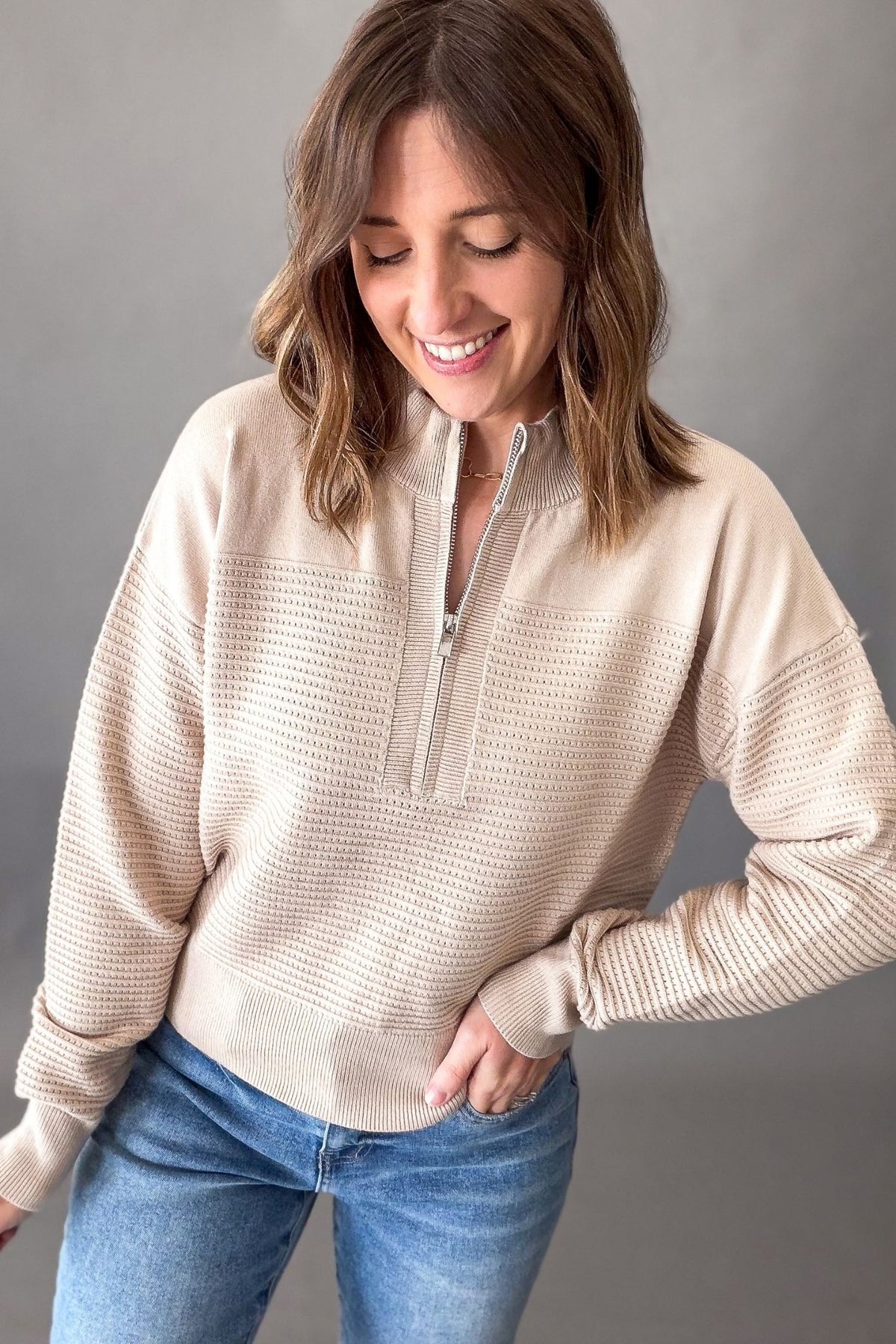 A woman wearing a beige textured quarter-zip pullover sweater and blue jeans, standing against a neutral gray background.