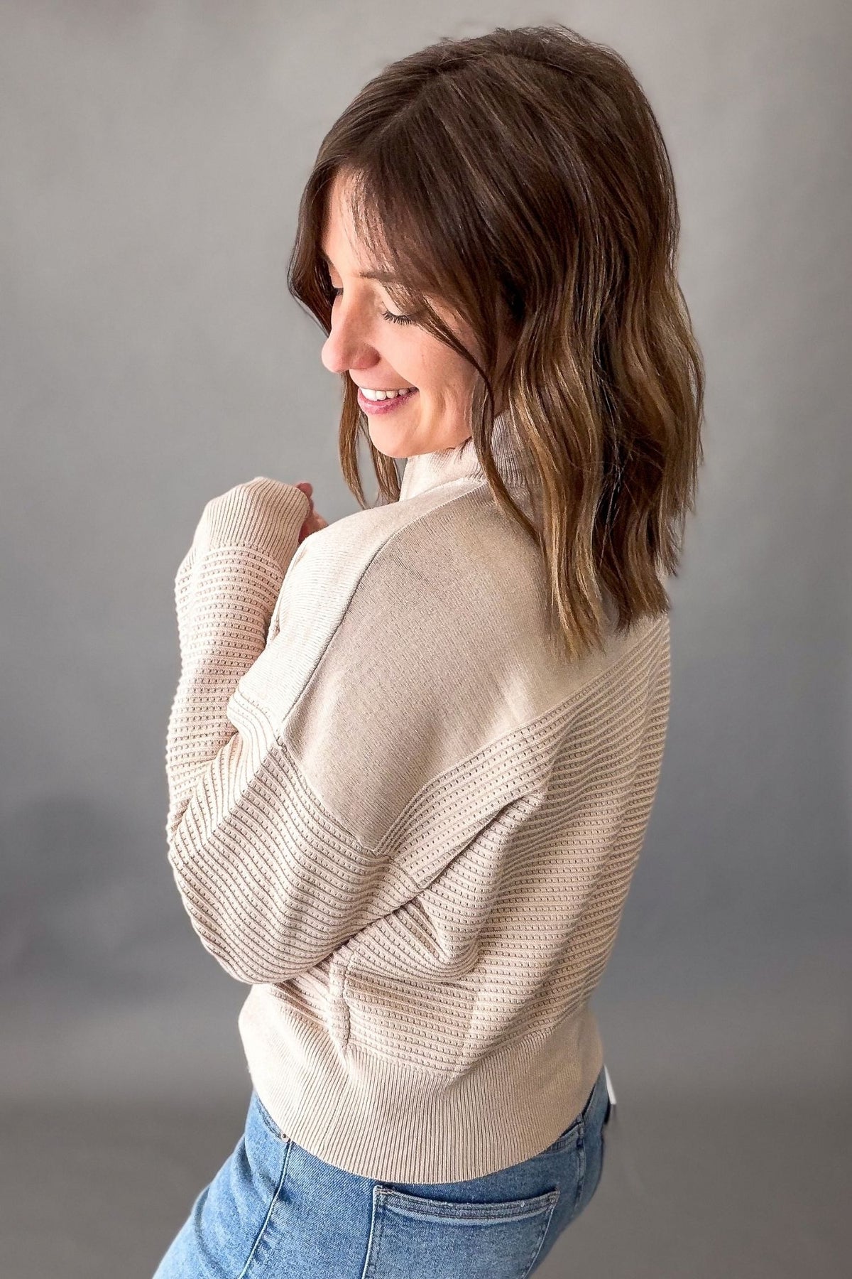 A woman wearing a beige ribbed knit mock neck sweater and blue jeans, photographed from the back against a neutral gray studio background.
