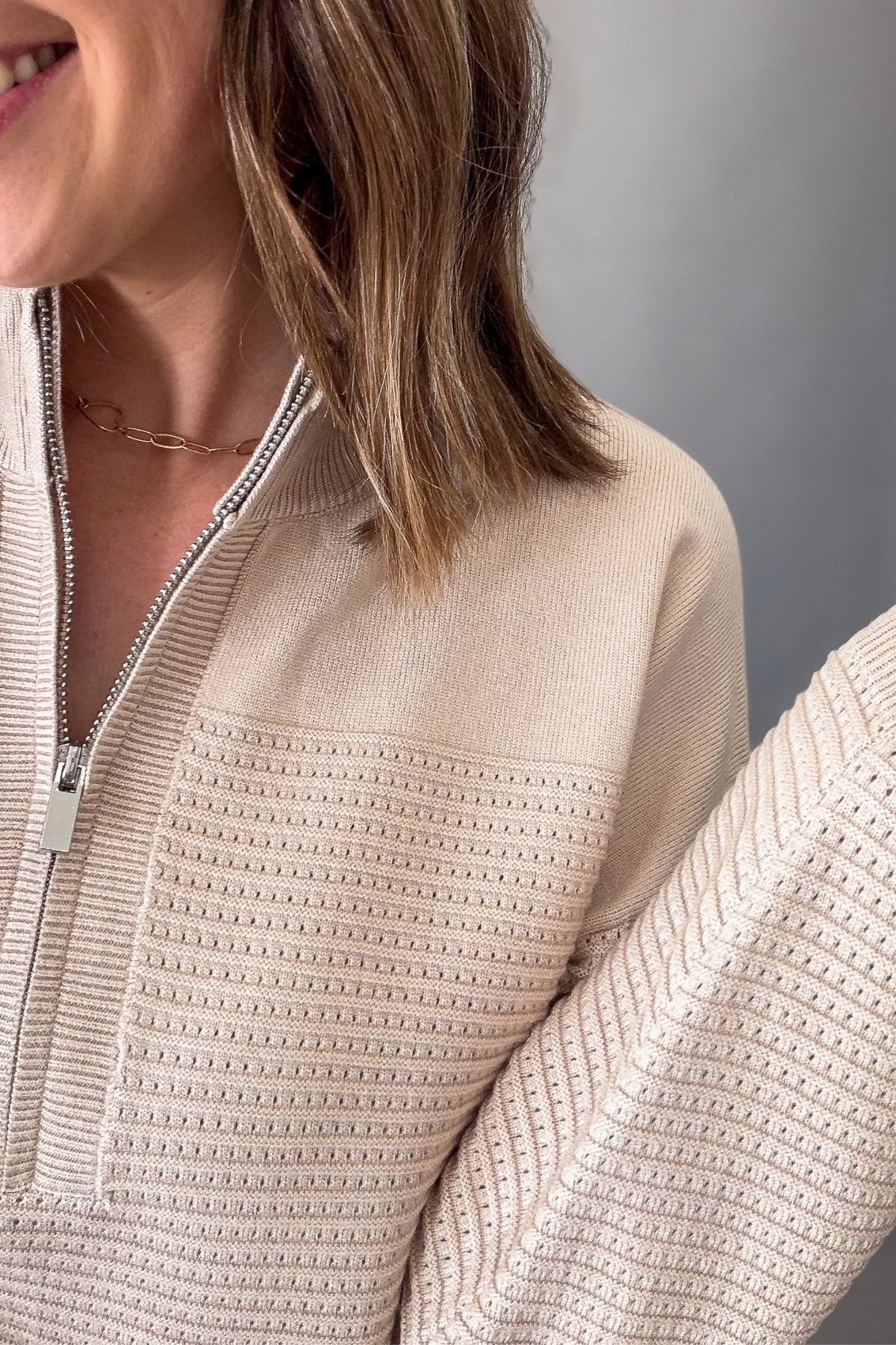 Close-up of a woman wearing a textured knit quarter-zip sweater in a warm cream or oatmeal color, showing the ribbed fabric, zipper, and part of her hair and skin.